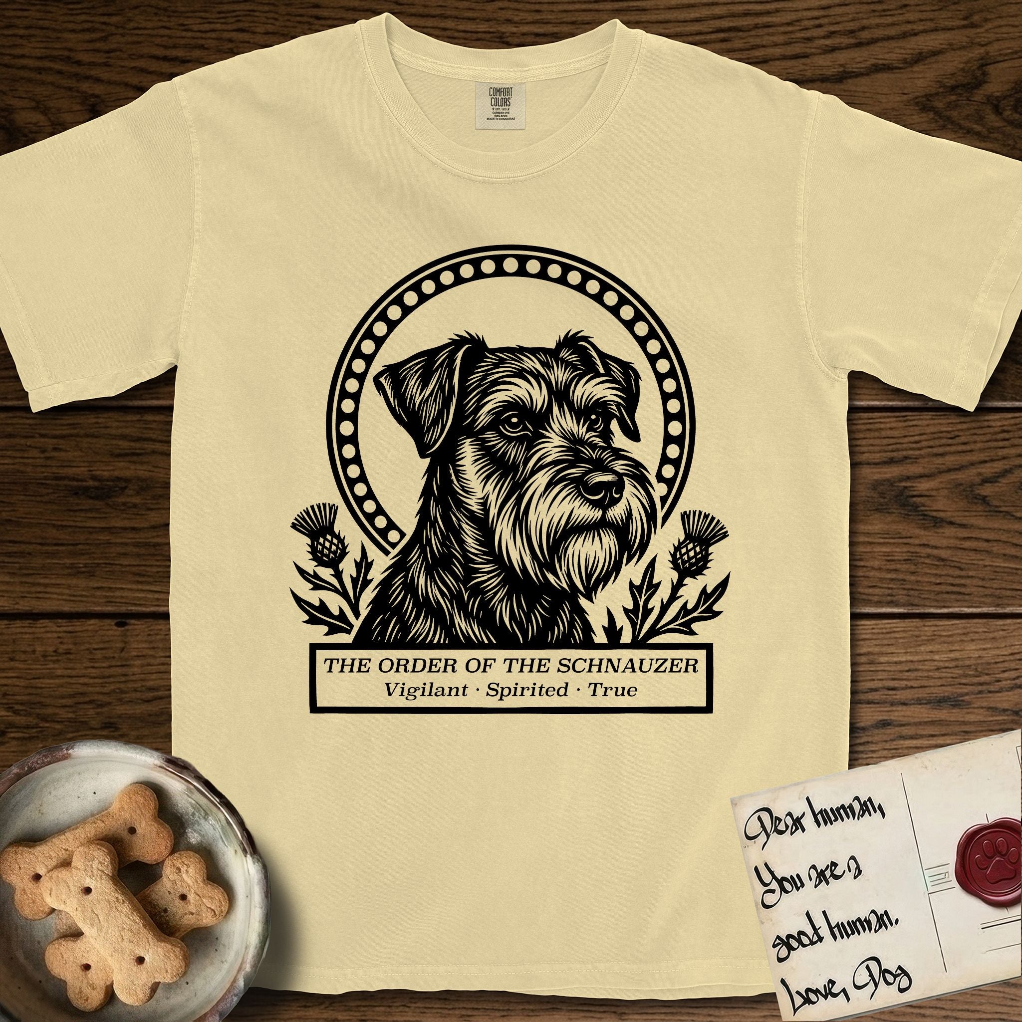 Holy Orders Schnauzer T-Shirt flat lay in Butter, featuring a bold black-and-white linocut saint-medallion portrait of a Schnauzer.