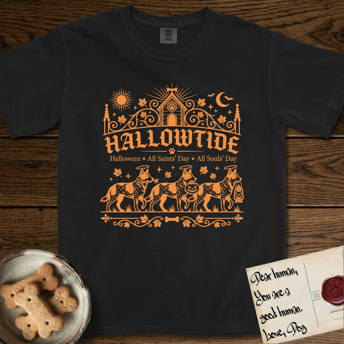 Hallowtide Australian Cattle Dog T-Shirt flat lay in Black, featuring Orange one-color gothic illustration of three haloed speckled Australian Cattle Dogs walking in procession carrying a skeleton key, a scary jack-o'-lantern, and an iron lantern, under the blackletter headline HALLOWTIDE.