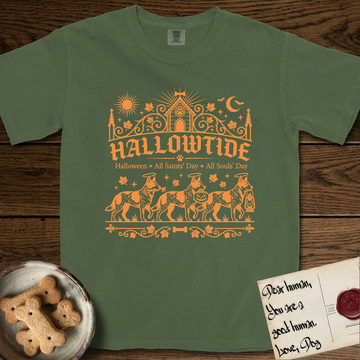 Hallowtide Australian Cattle Dog T-Shirt flat lay in Moss, featuring Orange one-color gothic illustration of three haloed speckled Australian Cattle Dogs walking in procession carrying a skeleton key, a scary jack-o'-lantern, and an iron lantern, under the blackletter headline HALLOWTIDE.