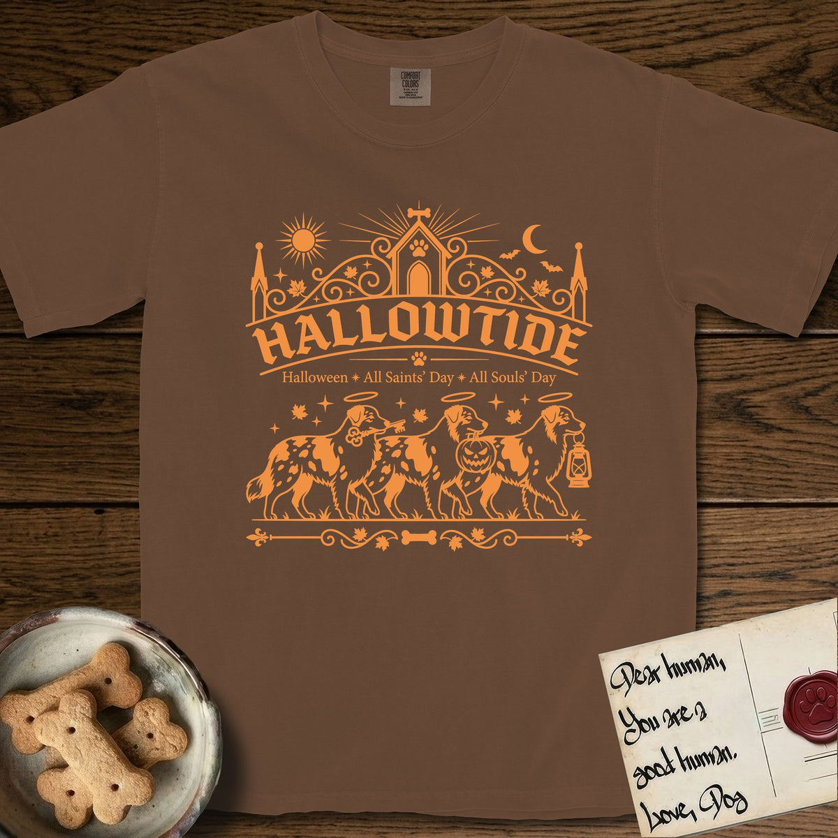 Hallowtide Australian Shepherd T-Shirt flat lay in Espresso, featuring Orange one-color gothic illustration of three haloed merle Australian Shepherds walking in procession carrying a skeleton key, a scary jack-o'-lantern, and an iron lantern, under the blackletter headline HALLOWTIDE.