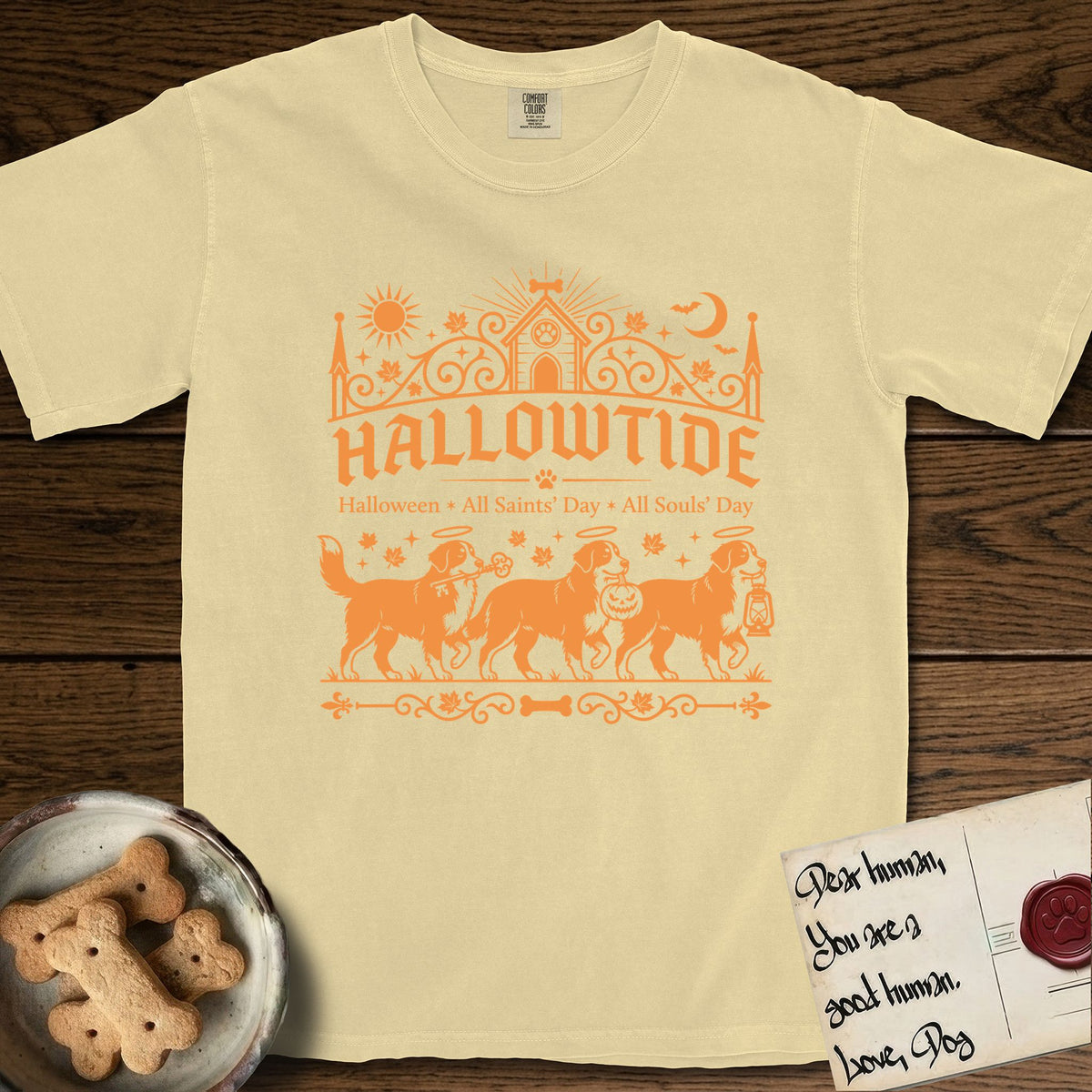 Hallowtide Bernese Mountain Dog T-Shirt flat lay in Butter, featuring Orange one-color gothic illustration of three haloed Bernese Mountain Dogs walking in procession carrying a skeleton key, a scary jack-o'-lantern, and an iron lantern, under the blackletter headline HALLOWTIDE.