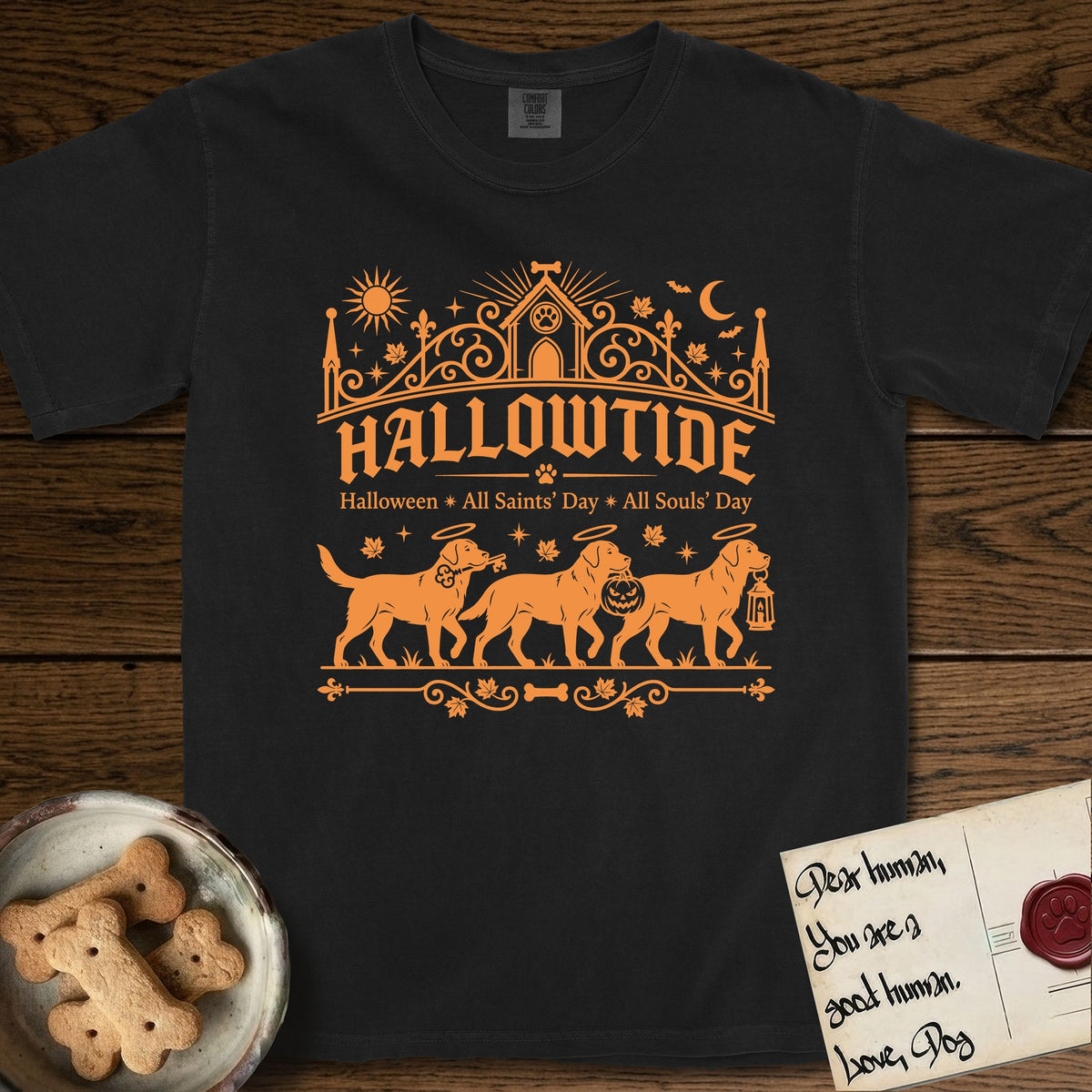 Hallowtide Black Lab T-Shirt flat lay in Black, featuring Orange one-color gothic illustration of three haloed Black Labs walking in procession carrying a skeleton key, a scary jack-o'-lantern, and an iron lantern, under the blackletter headline HALLOWTIDE.