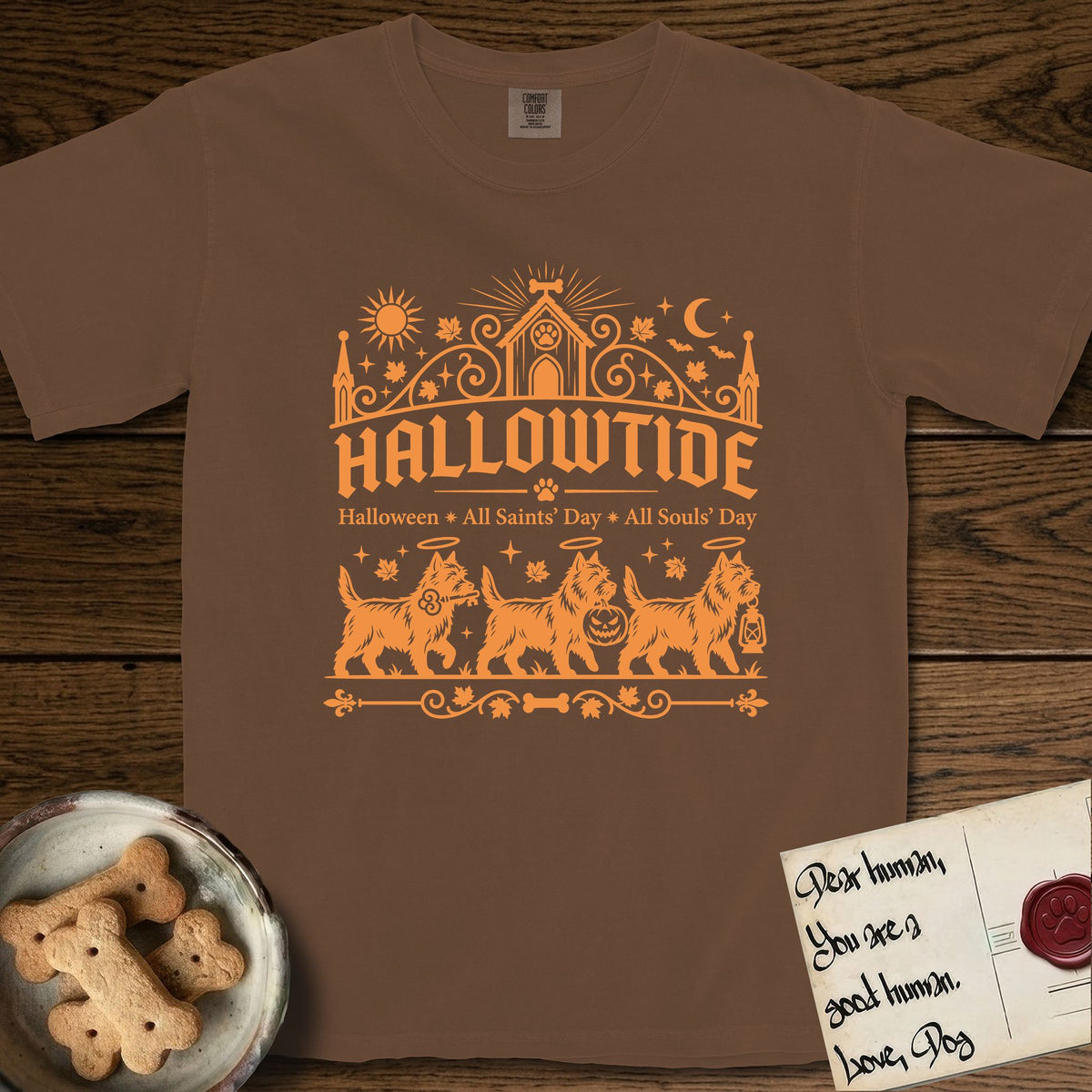 Hallowtide Cairn Terrier T-Shirt flat lay in Espresso, featuring Orange one-color gothic illustration of three haloed shaggy Cairn Terriers walking in procession carrying a skeleton key, a scary jack-o'-lantern, and an iron lantern, under the blackletter headline HALLOWTIDE.