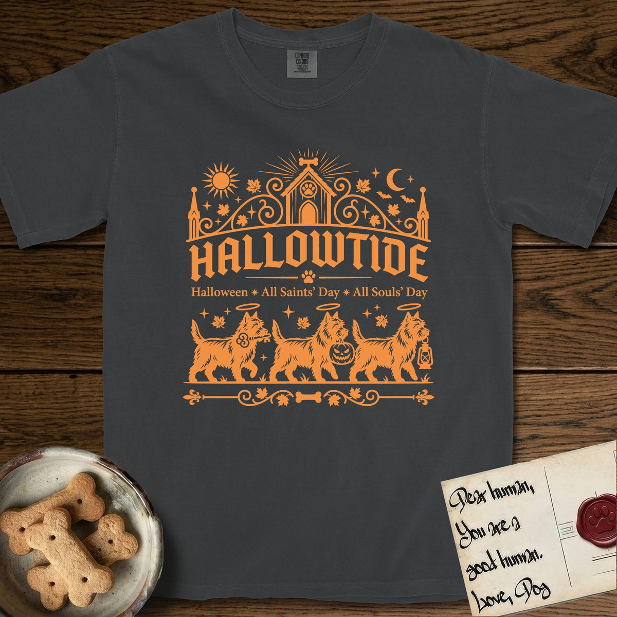 Hallowtide Cairn Terrier T-Shirt flat lay in Graphite, featuring Orange one-color gothic illustration of three haloed shaggy Cairn Terriers walking in procession carrying a skeleton key, a scary jack-o'-lantern, and an iron lantern, under the blackletter headline HALLOWTIDE.