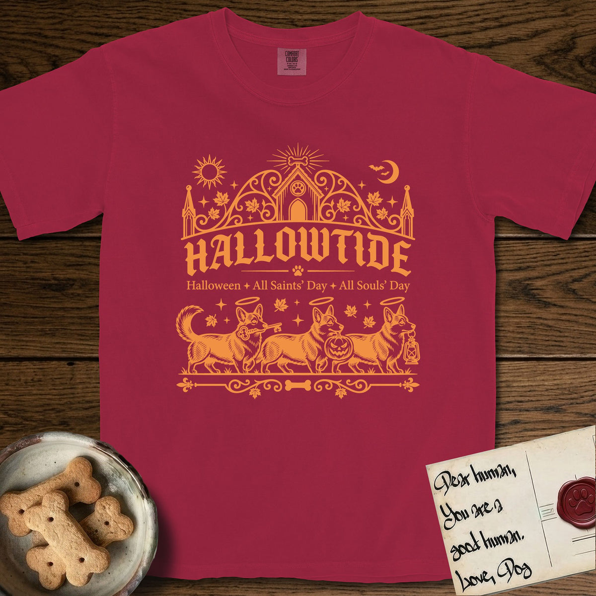 Hallowtide Corgi T-Shirt flat lay in Chili, featuring Orange one-color gothic illustration of three haloed Corgis walking in procession carrying a skeleton key, a scary jack-o'-lantern, and an iron lantern, under the blackletter headline HALLOWTIDE.