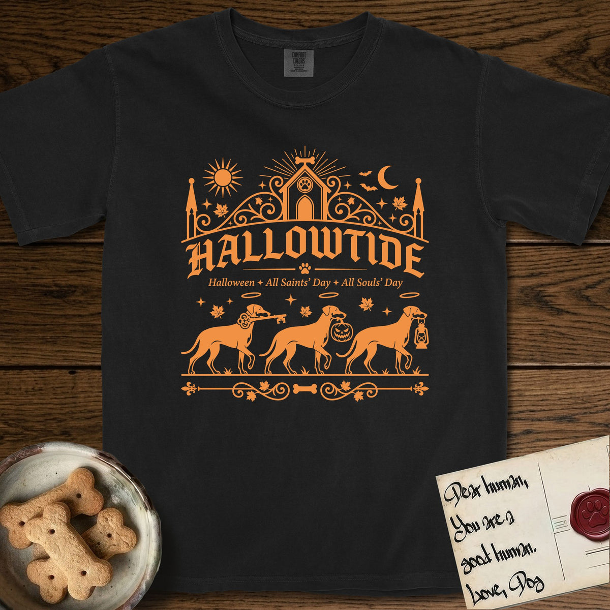 Hallowtide Doberman T-Shirt flat lay in Black, featuring Orange one-color gothic illustration of three haloed Dobermans walking in procession carrying a skeleton key, a scary jack-o'-lantern, and an iron lantern, under the blackletter headline HALLOWTIDE.
