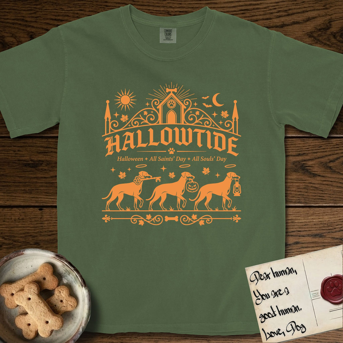 Hallowtide Doberman T-Shirt flat lay in Moss, featuring Orange one-color gothic illustration of three haloed Dobermans walking in procession carrying a skeleton key, a scary jack-o'-lantern, and an iron lantern, under the blackletter headline HALLOWTIDE.