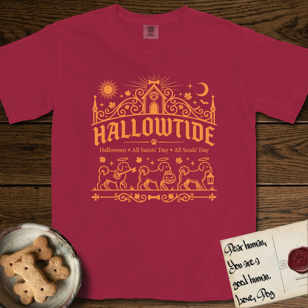 Hallowtide Maltese T-Shirt flat lay in Chili, featuring Orange one-color gothic illustration of three haloed Maltese walking in procession carrying a skeleton key, a scary jack-o'-lantern, and an iron lantern, under the blackletter headline HALLOWTIDE.
