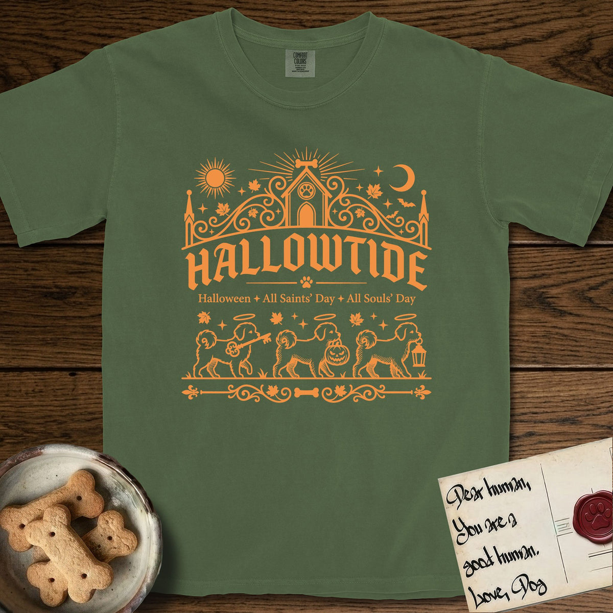 Hallowtide Maltese T-Shirt flat lay in Moss, featuring Orange one-color gothic illustration of three haloed Maltese walking in procession carrying a skeleton key, a scary jack-o'-lantern, and an iron lantern, under the blackletter headline HALLOWTIDE.