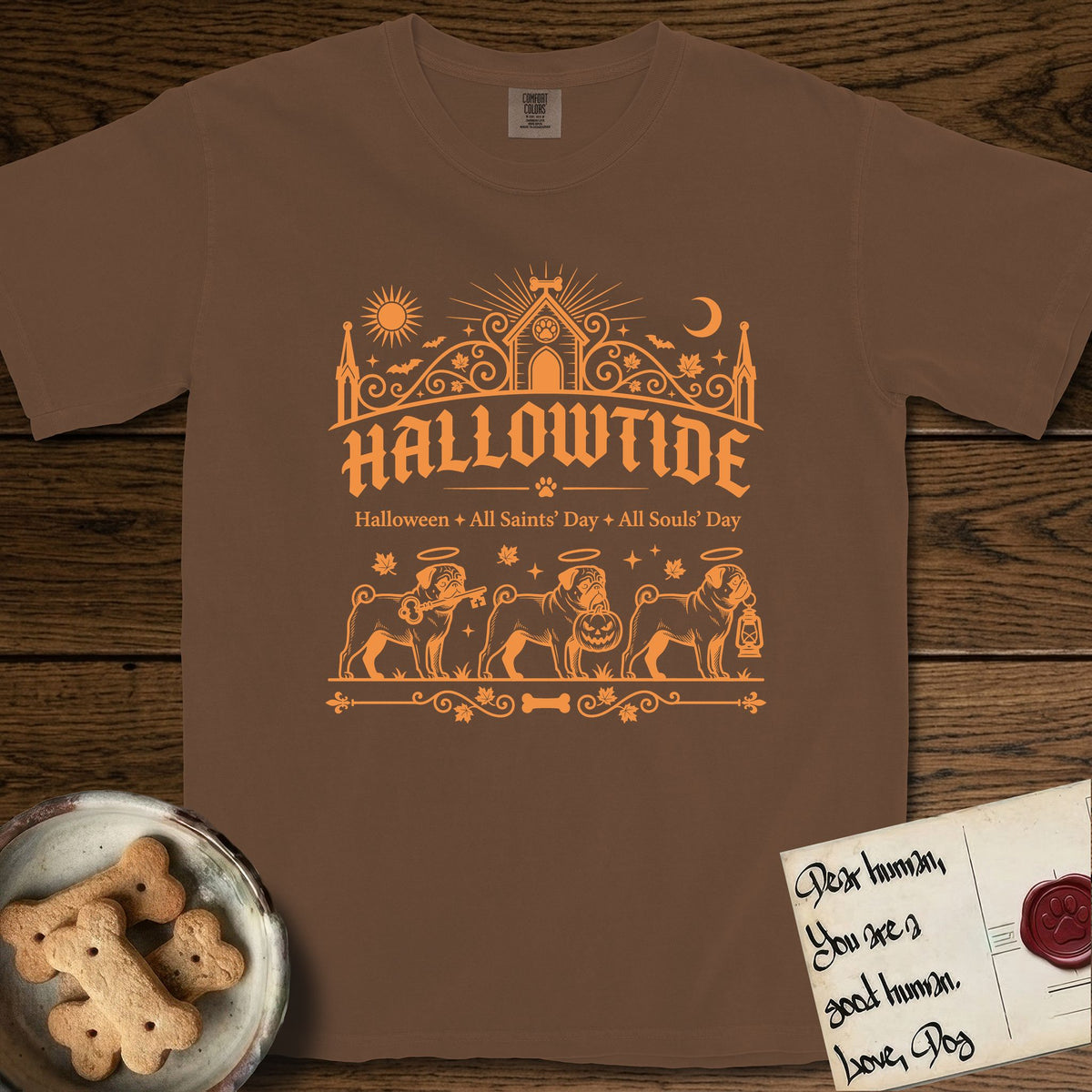 Hallowtide Pug T-Shirt flat lay in Espresso, featuring Orange one-color gothic illustration of three haloed curly-tailed Pugs walking in procession carrying a skeleton key, a scary jack-o'-lantern, and an iron lantern, under the blackletter headline HALLOWTIDE.