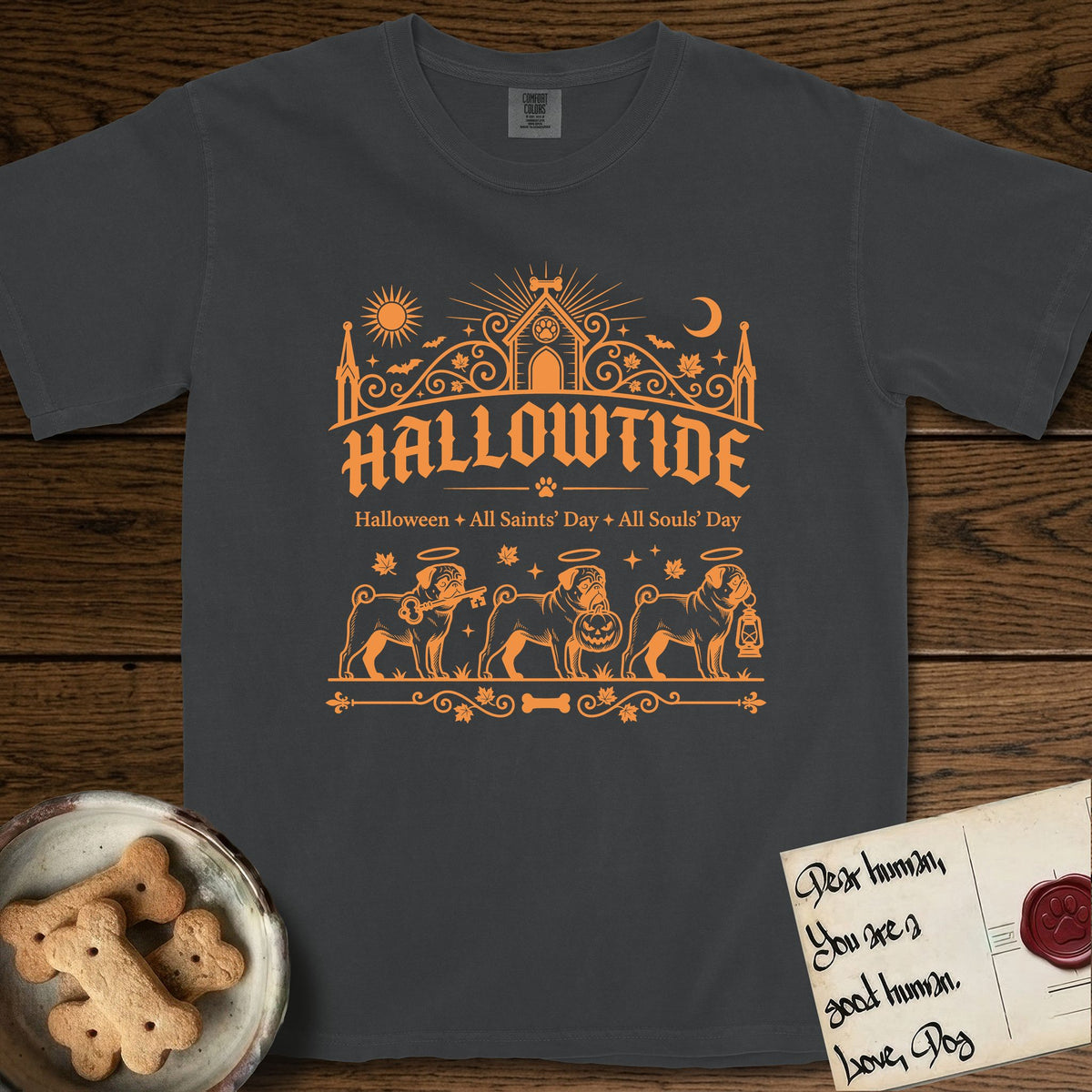 Hallowtide Pug T-Shirt flat lay in Graphite, featuring Orange one-color gothic illustration of three haloed curly-tailed Pugs walking in procession carrying a skeleton key, a scary jack-o'-lantern, and an iron lantern, under the blackletter headline HALLOWTIDE.
