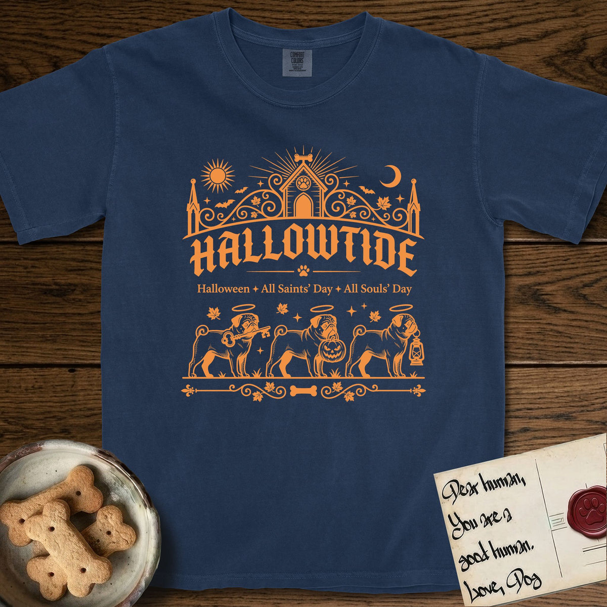 Hallowtide Pug T-Shirt flat lay in True Navy, featuring Orange one-color gothic illustration of three haloed curly-tailed Pugs walking in procession carrying a skeleton key, a scary jack-o'-lantern, and an iron lantern, under the blackletter headline HALLOWTIDE.