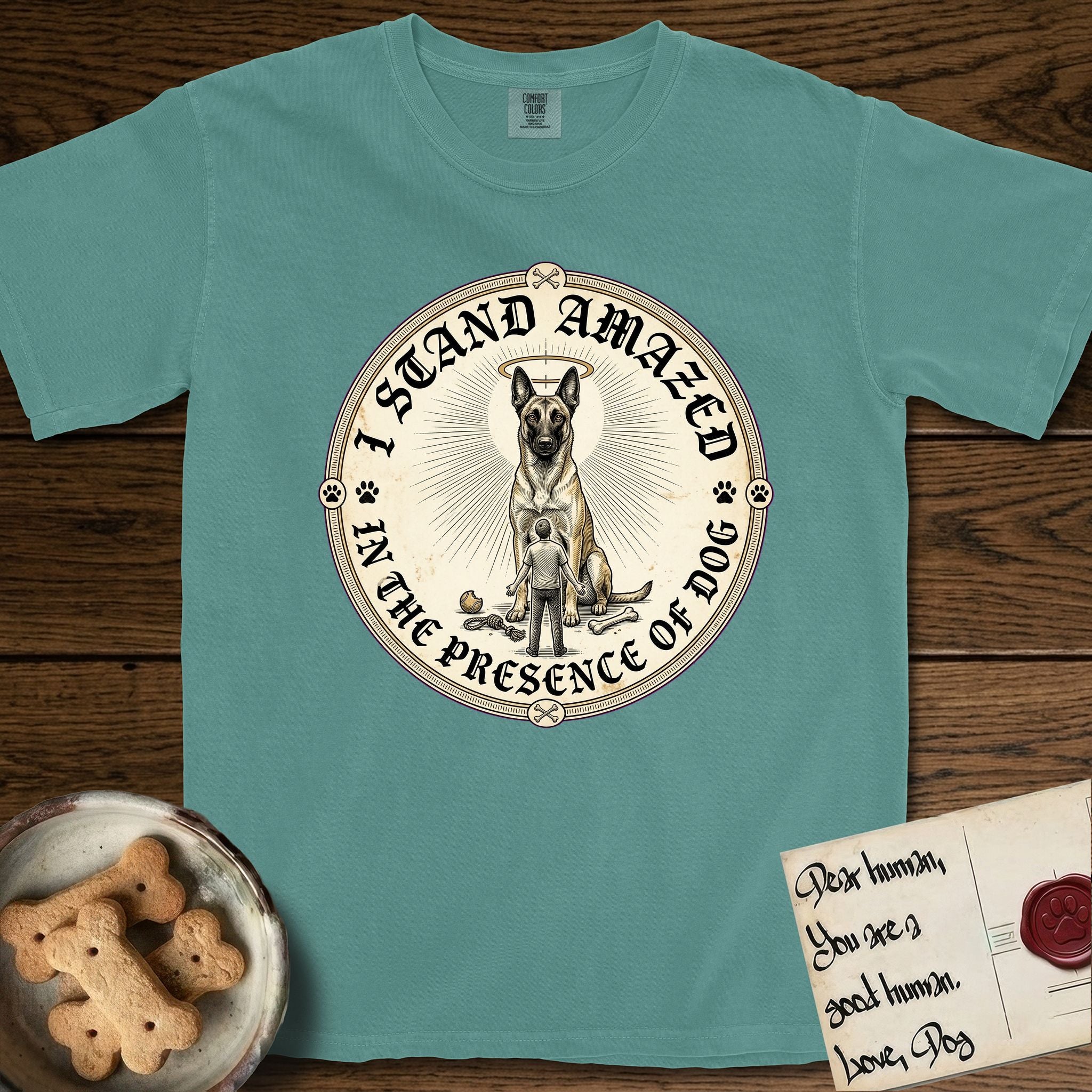 I Stand Amazed in the Presence Belgian Malinois T-Shirt flat lay in Blue Spruce, featuring a vintage engraving medallion of a haloed Belgian Malinois with a small devoted human gazing up from below.