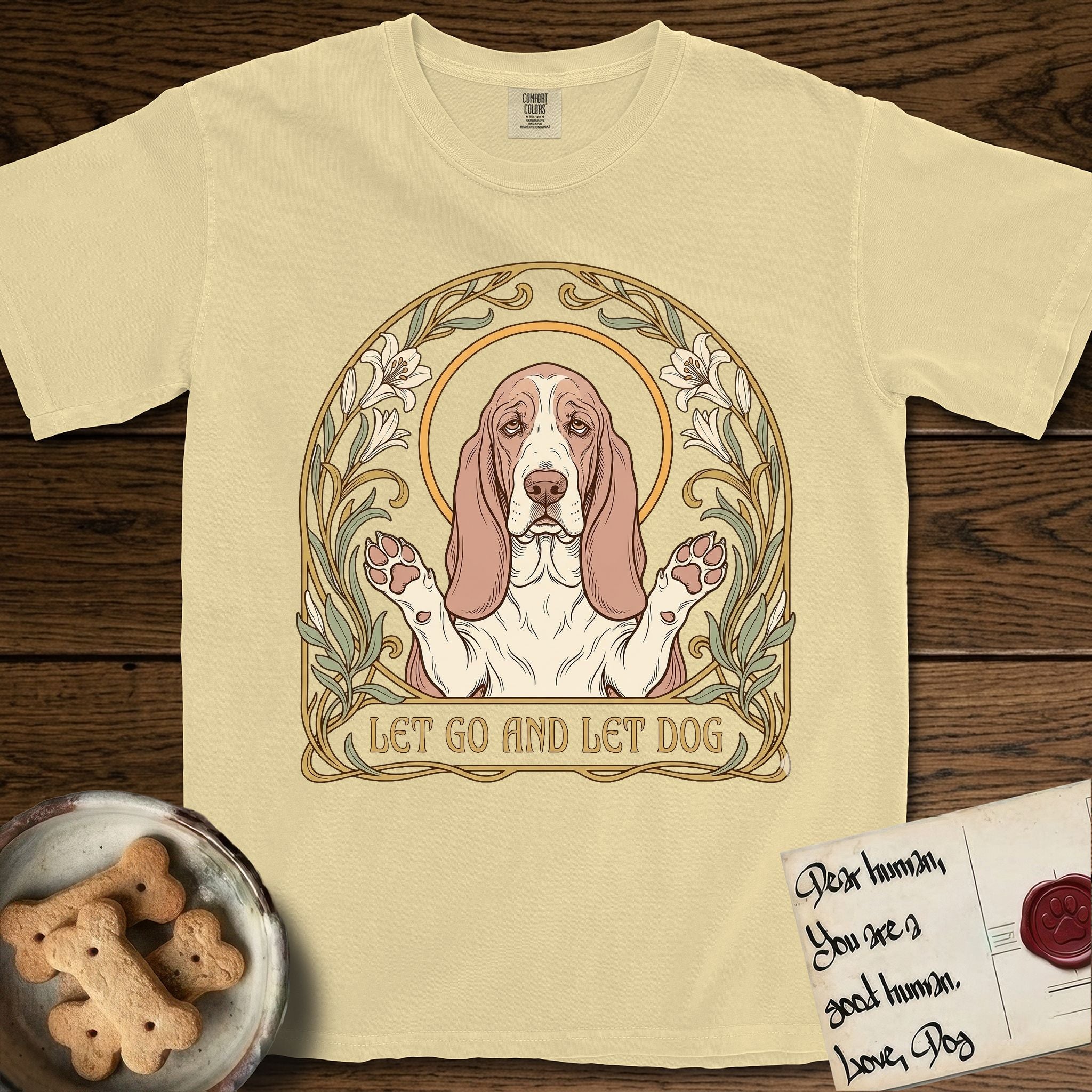 Let Go and Let Dog Basset Hound T-Shirt flat lay in Butter, featuring an Art Nouveau botanical illustration of a haloed Basset Hound with paws raised in blessing.