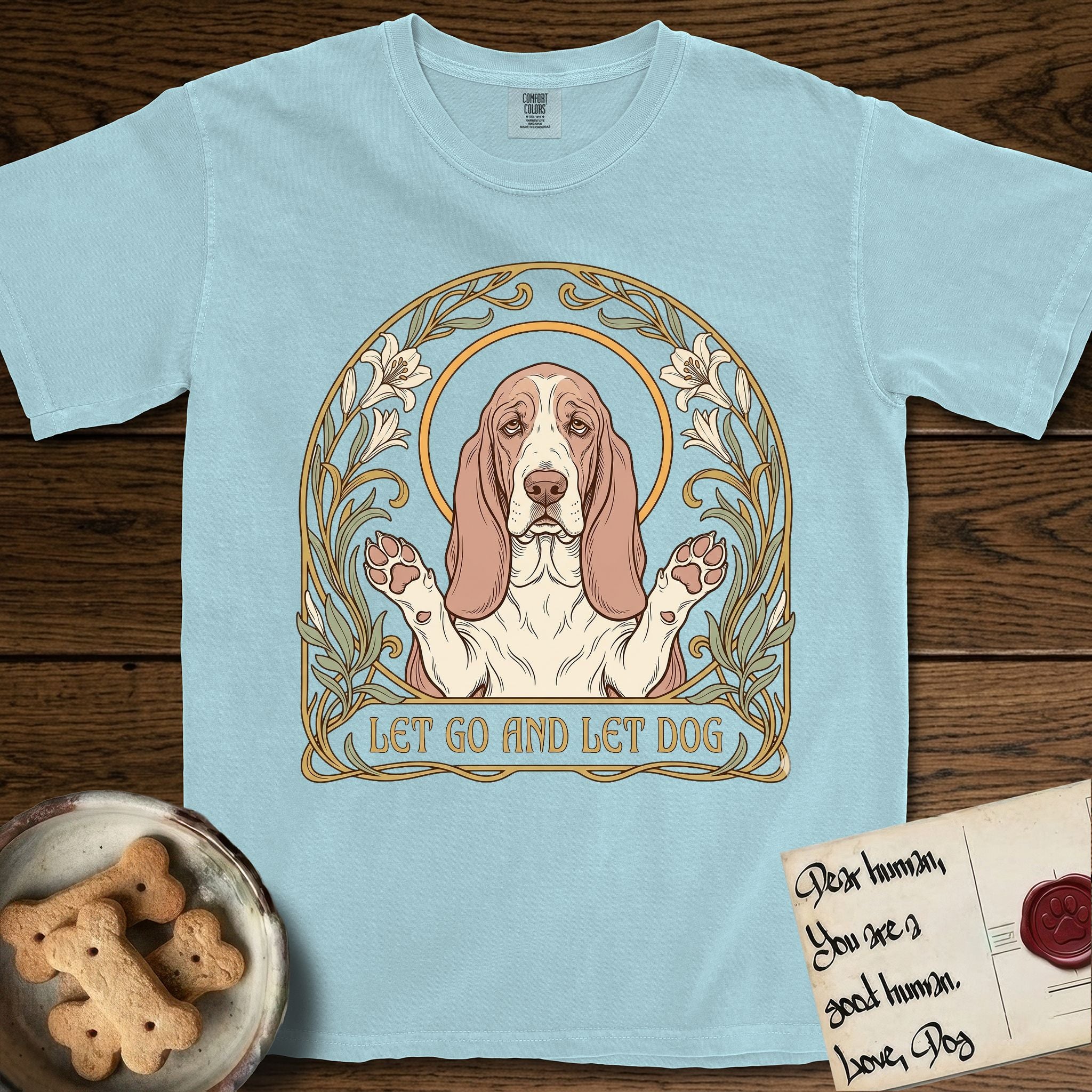 Let Go and Let Dog Basset Hound T-Shirt flat lay in Chambray, featuring an Art Nouveau botanical illustration of a haloed Basset Hound with paws raised in blessing.
