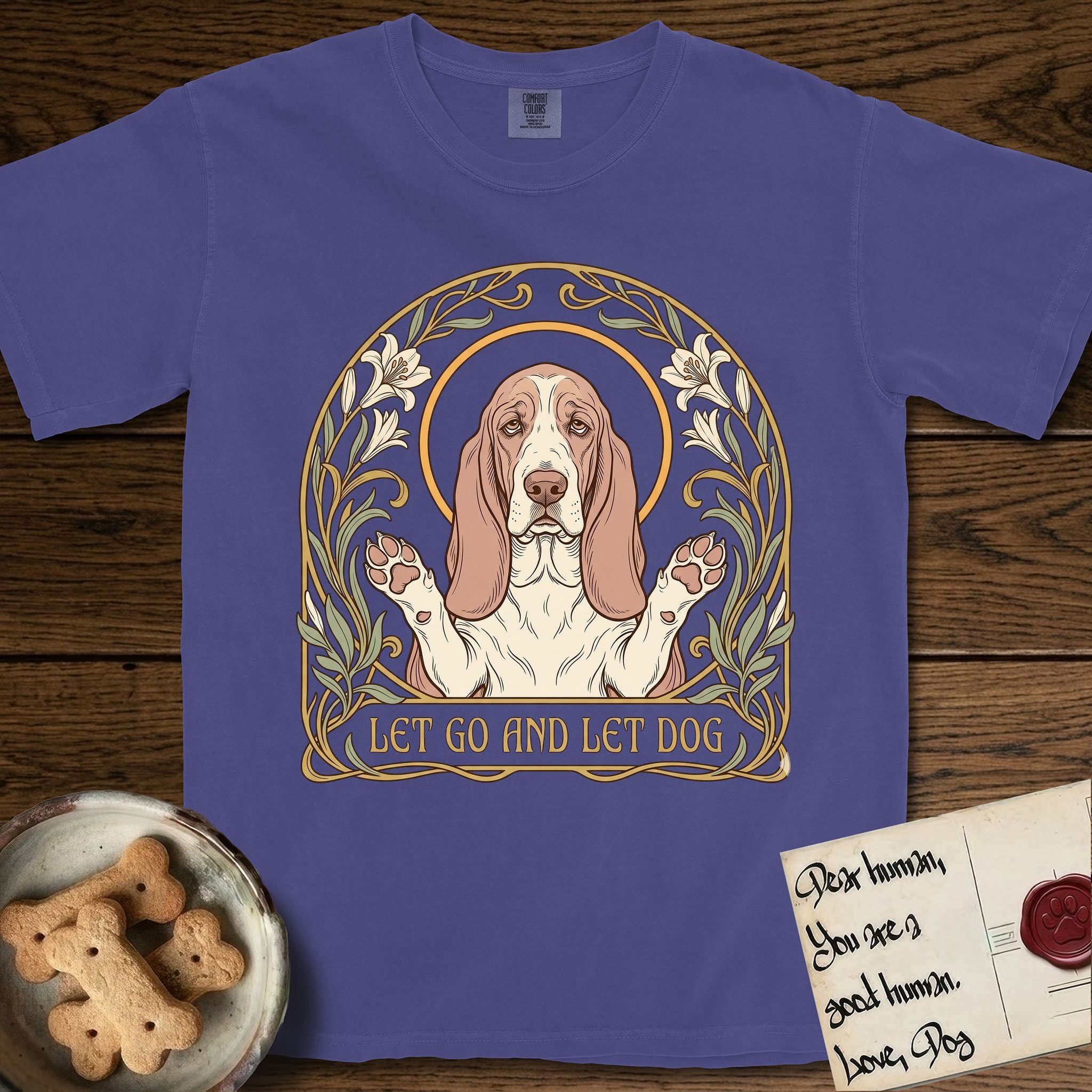 Let Go and Let Dog Basset Hound T-Shirt flat lay in Grape, featuring an Art Nouveau botanical illustration of a haloed Basset Hound with paws raised in blessing.