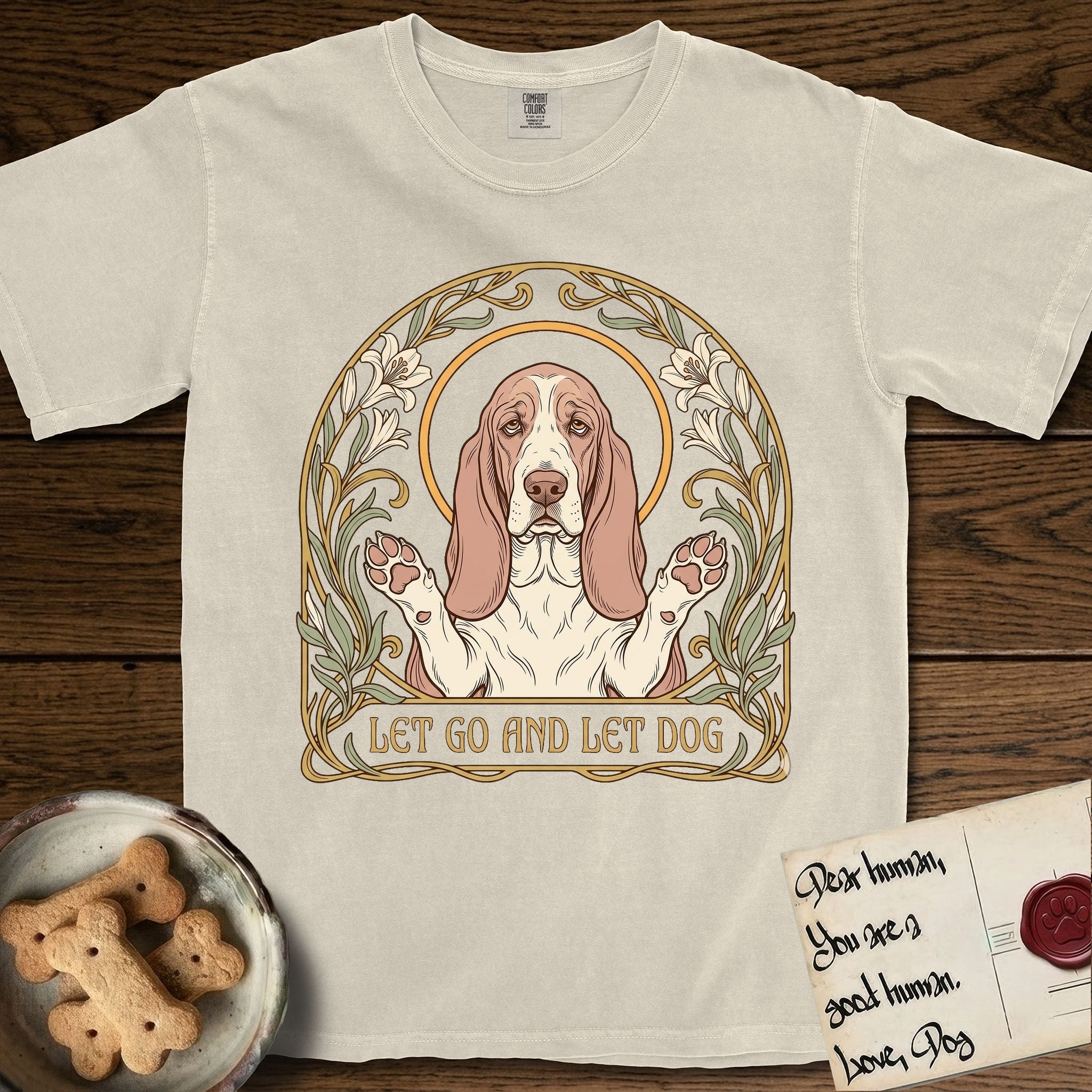 Let Go and Let Dog Basset Hound T-Shirt flat lay in Ivory, featuring an Art Nouveau botanical illustration of a haloed Basset Hound with paws raised in blessing.