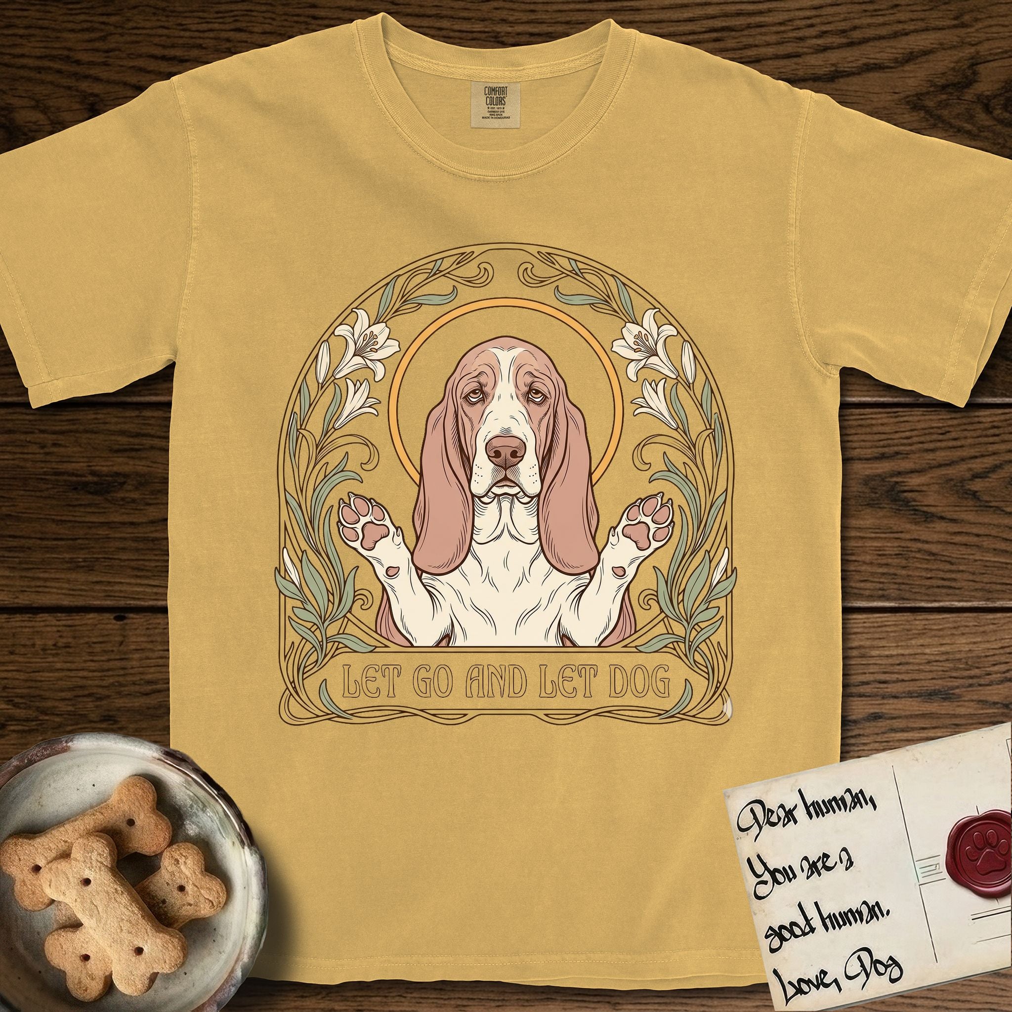 Let Go and Let Dog Basset Hound T-Shirt flat lay in Mustard, featuring an Art Nouveau botanical illustration of a haloed Basset Hound with paws raised in blessing.