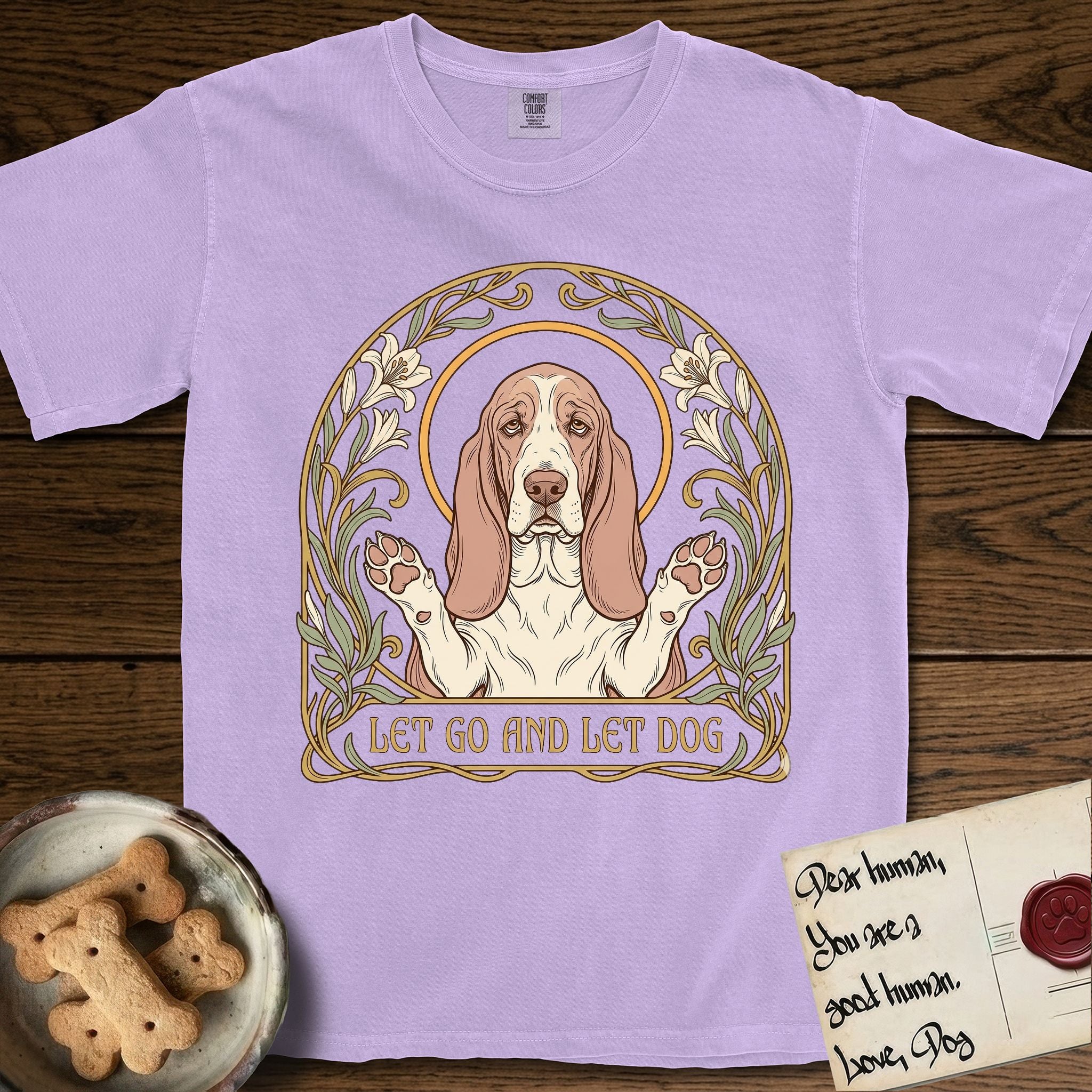 Let Go and Let Dog Basset Hound T-Shirt flat lay in Orchid, featuring an Art Nouveau botanical illustration of a haloed Basset Hound with paws raised in blessing.