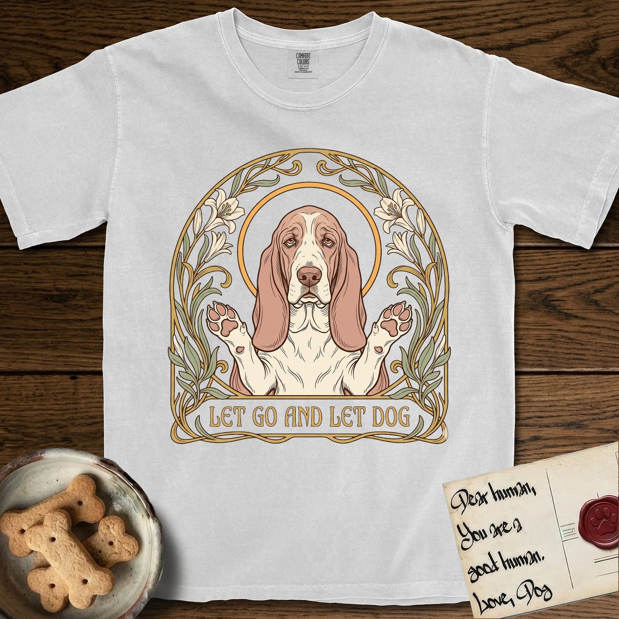 Let Go and Let Dog Basset Hound T-Shirt flat lay in White, featuring an Art Nouveau botanical illustration of a haloed Basset Hound with paws raised in blessing.