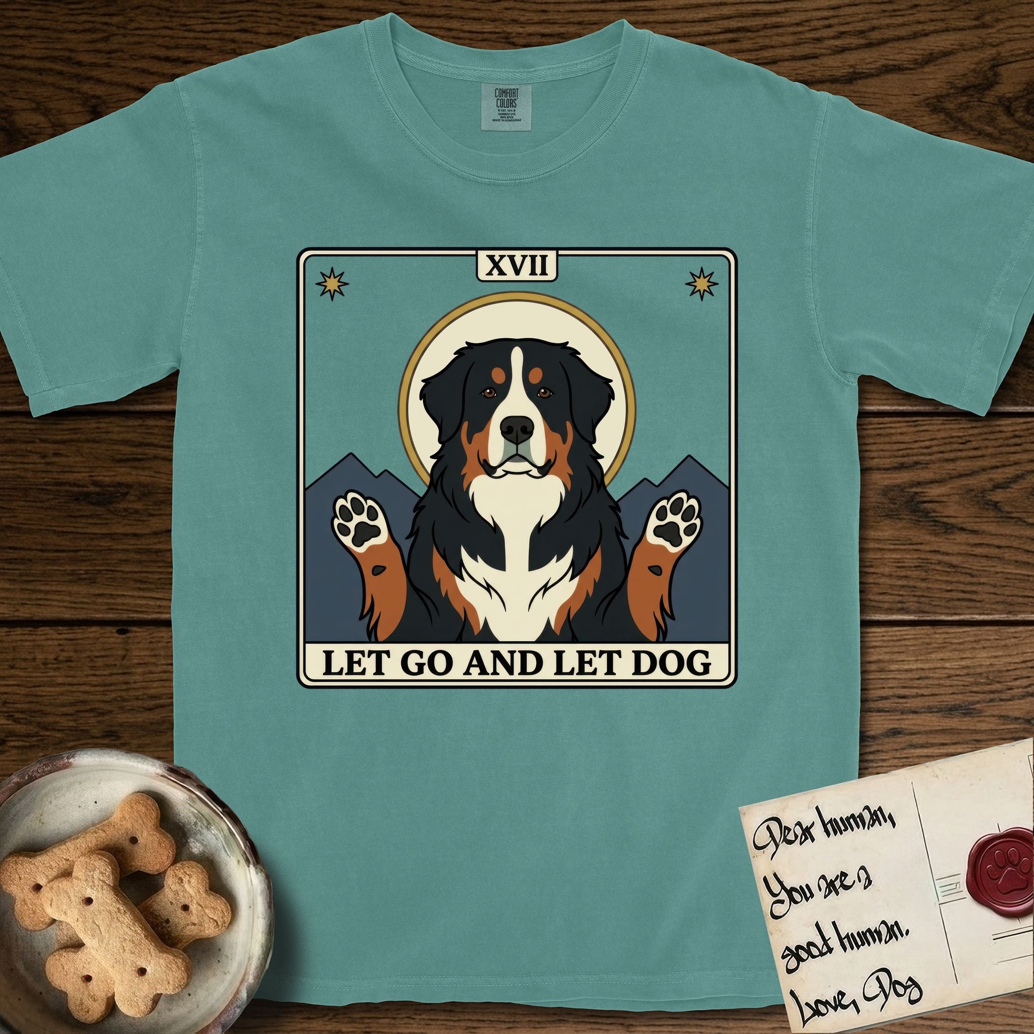 Let Go and Let Dog Bernese Mountain Dog T-Shirt flat lay in Blue Spruce, featuring a vintage tarot card with a haloed tricolor Bernese Mountain Dog, paws raised, mountains rising behind her shoulders.