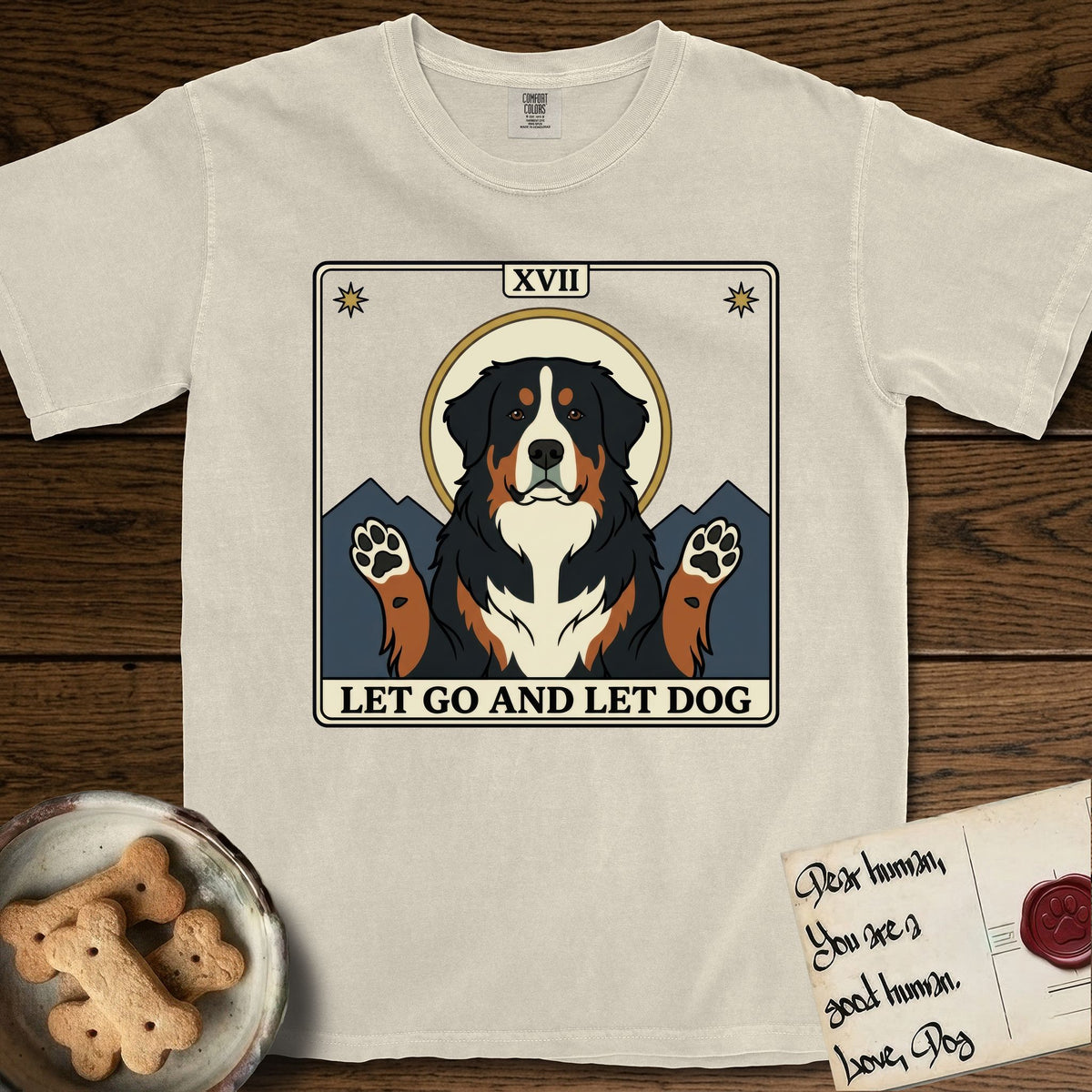 Let Go and Let Dog Bernese Mountain Dog T-Shirt flat lay in Ivory, featuring a vintage tarot card with a haloed tricolor Bernese Mountain Dog, paws raised, mountains rising behind her shoulders.