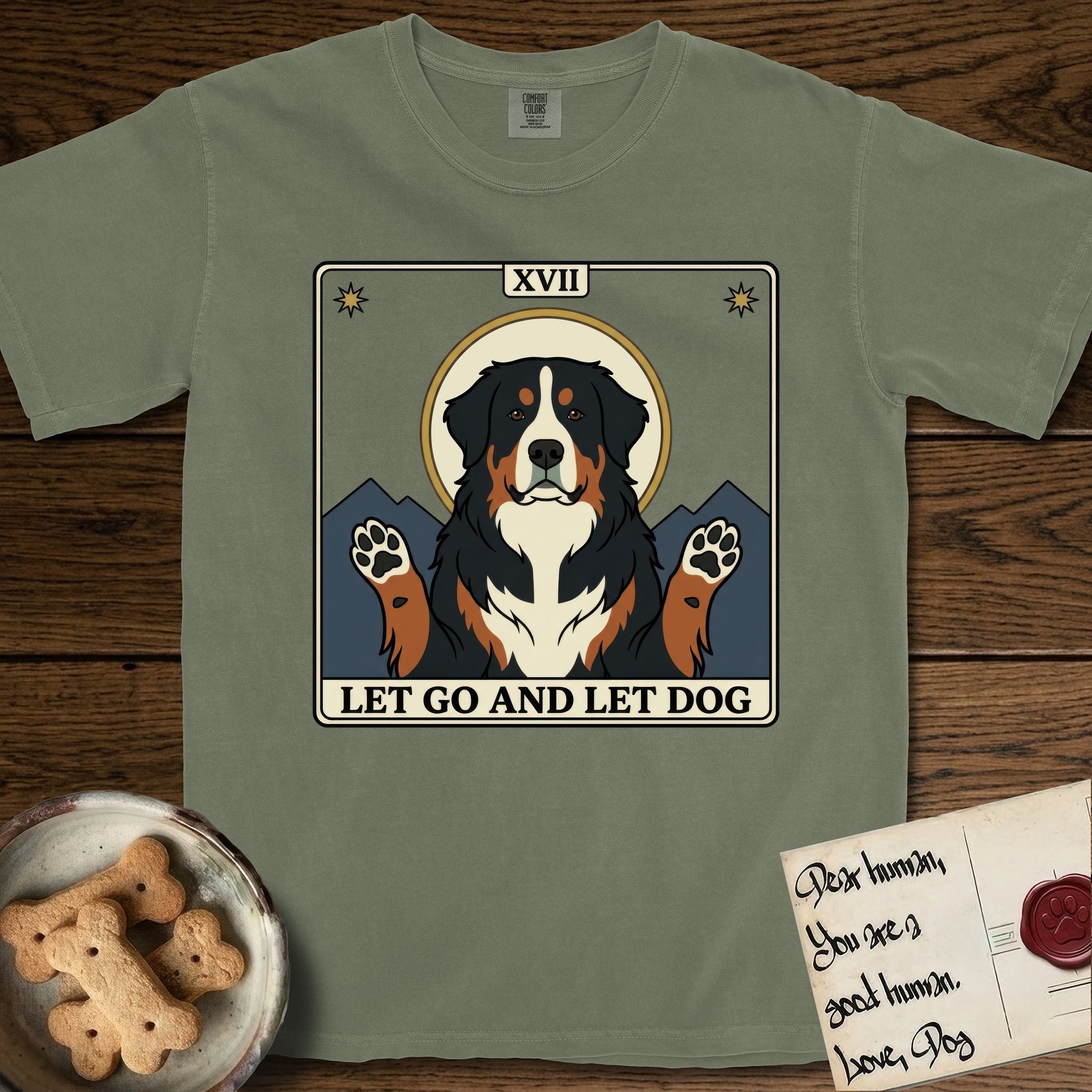 Let Go and Let Dog Bernese Mountain Dog T-Shirt flat lay in Sage, featuring a vintage tarot card with a haloed tricolor Bernese Mountain Dog, paws raised, mountains rising behind her shoulders.