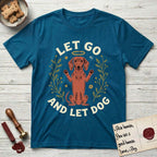 Let Go and Let Dog Dachshund T-Shirt flat lay in Antique Sapphire, featuring a folk-art botanical illustration of a haloed Dachshund sitting up with front paws raised.
