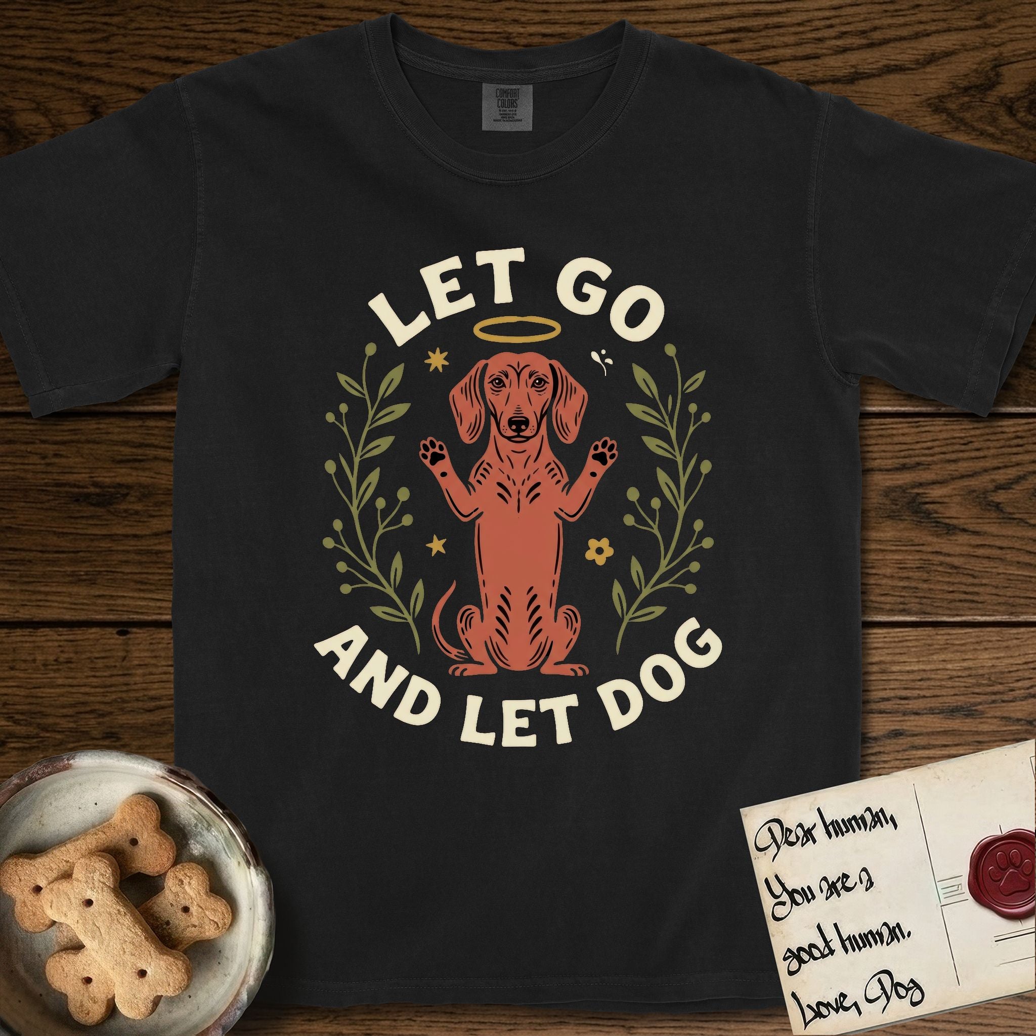 Let Go and Let Dog Dachshund T-Shirt flat lay in Black, featuring a folk-art botanical illustration of a haloed Dachshund sitting up with front paws raised.