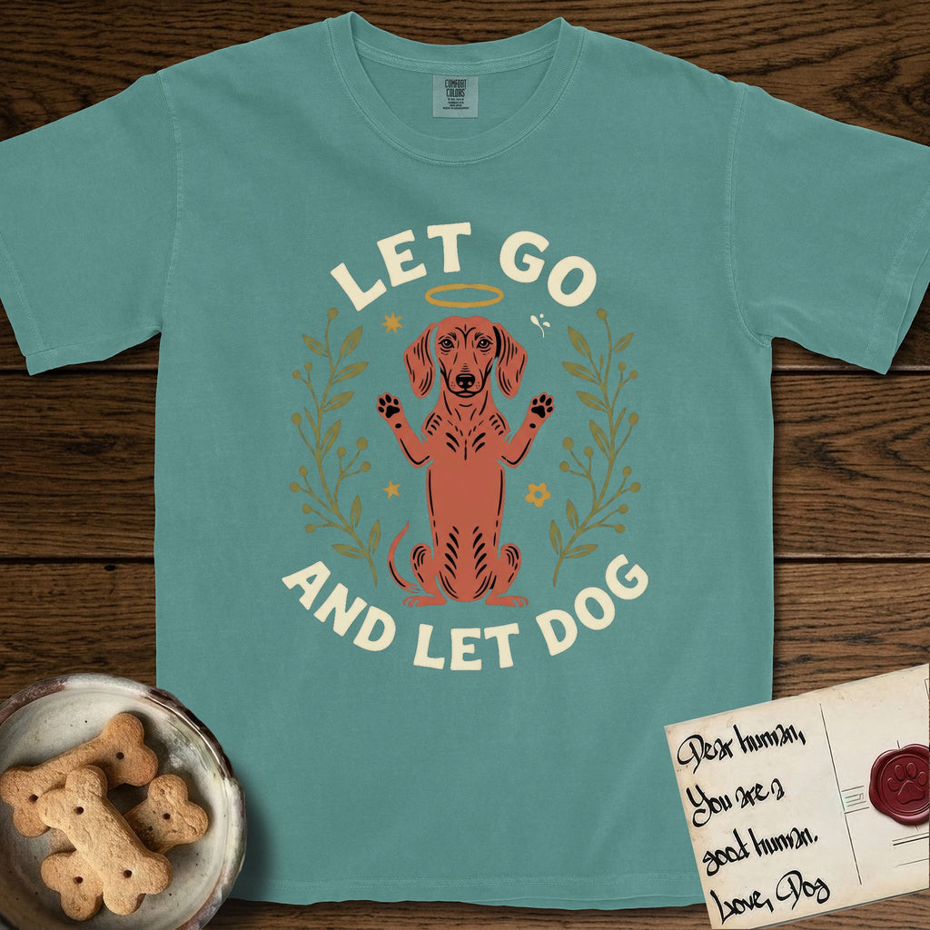 Let Go and Let Dog Dachshund T-Shirt flat lay in Blue Spruce, featuring a folk-art botanical illustration of a haloed Dachshund sitting up with front paws raised.