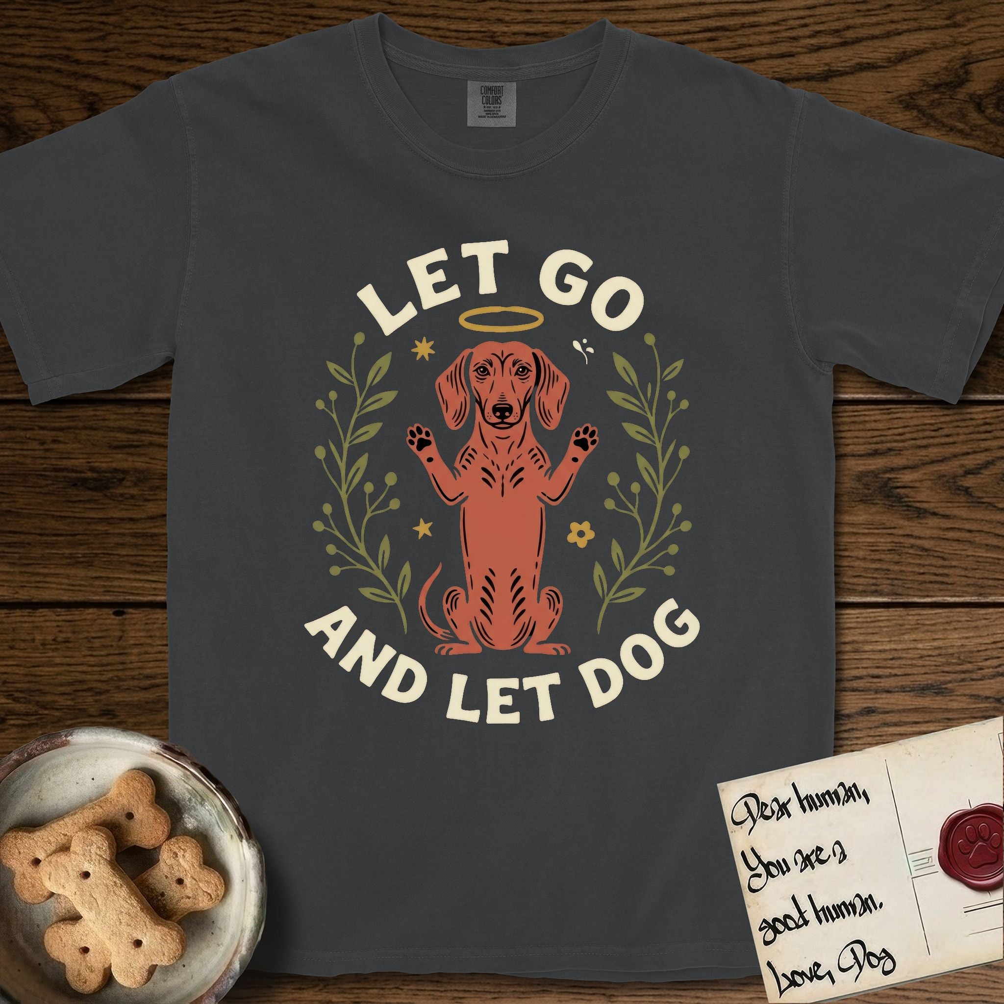 Let Go and Let Dog Dachshund T-Shirt flat lay in Graphite, featuring a folk-art botanical illustration of a haloed Dachshund sitting up with front paws raised.
