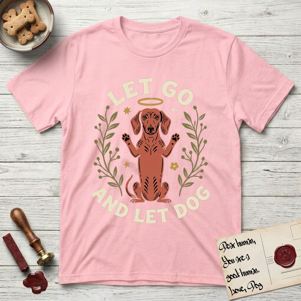 Let Go and Let Dog Dachshund T-Shirt flat lay in Light Pink, featuring a folk-art botanical illustration of a haloed Dachshund sitting up with front paws raised.