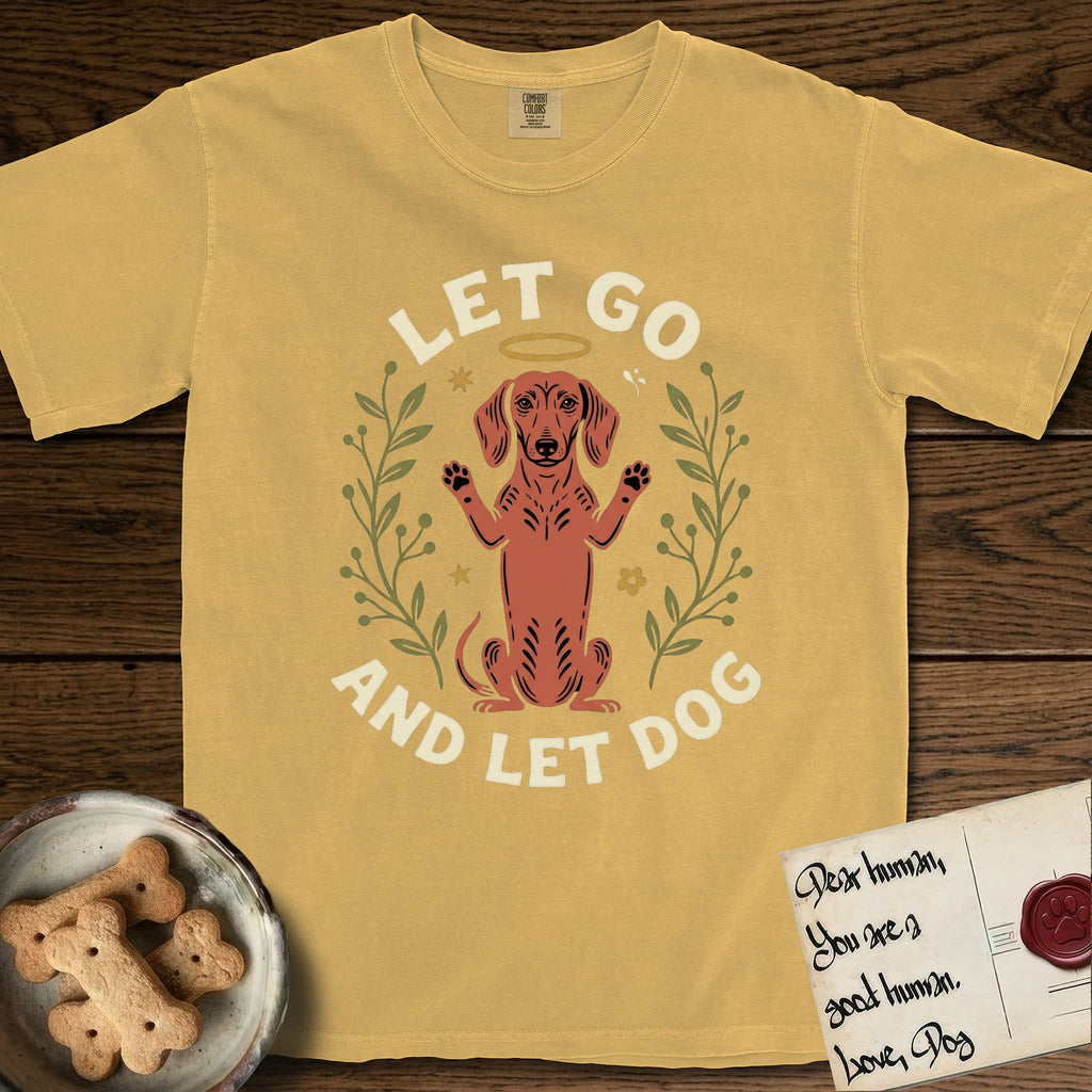 Let Go and Let Dog Dachshund T-Shirt flat lay in Mustard, featuring a folk-art botanical illustration of a haloed Dachshund sitting up with front paws raised.