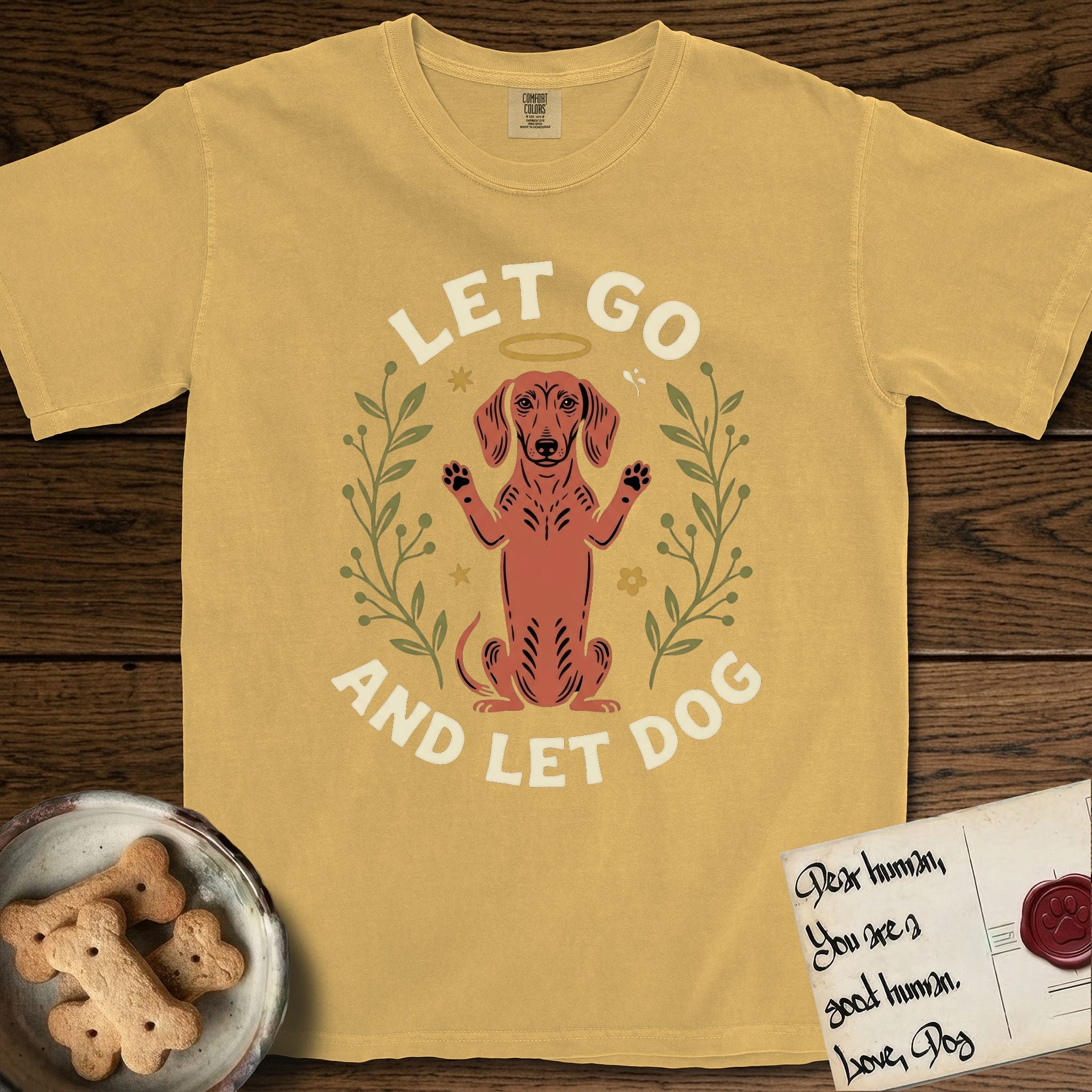Let Go and Let Dog Dachshund T-Shirt flat lay in Mustard, featuring a folk-art botanical illustration of a haloed Dachshund sitting up with front paws raised.