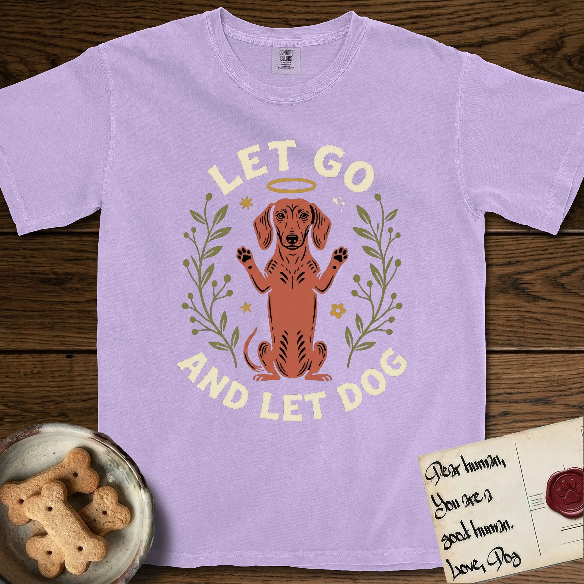 Let Go and Let Dog Dachshund T-Shirt flat lay in Orchid, featuring a folk-art botanical illustration of a haloed Dachshund sitting up with front paws raised.