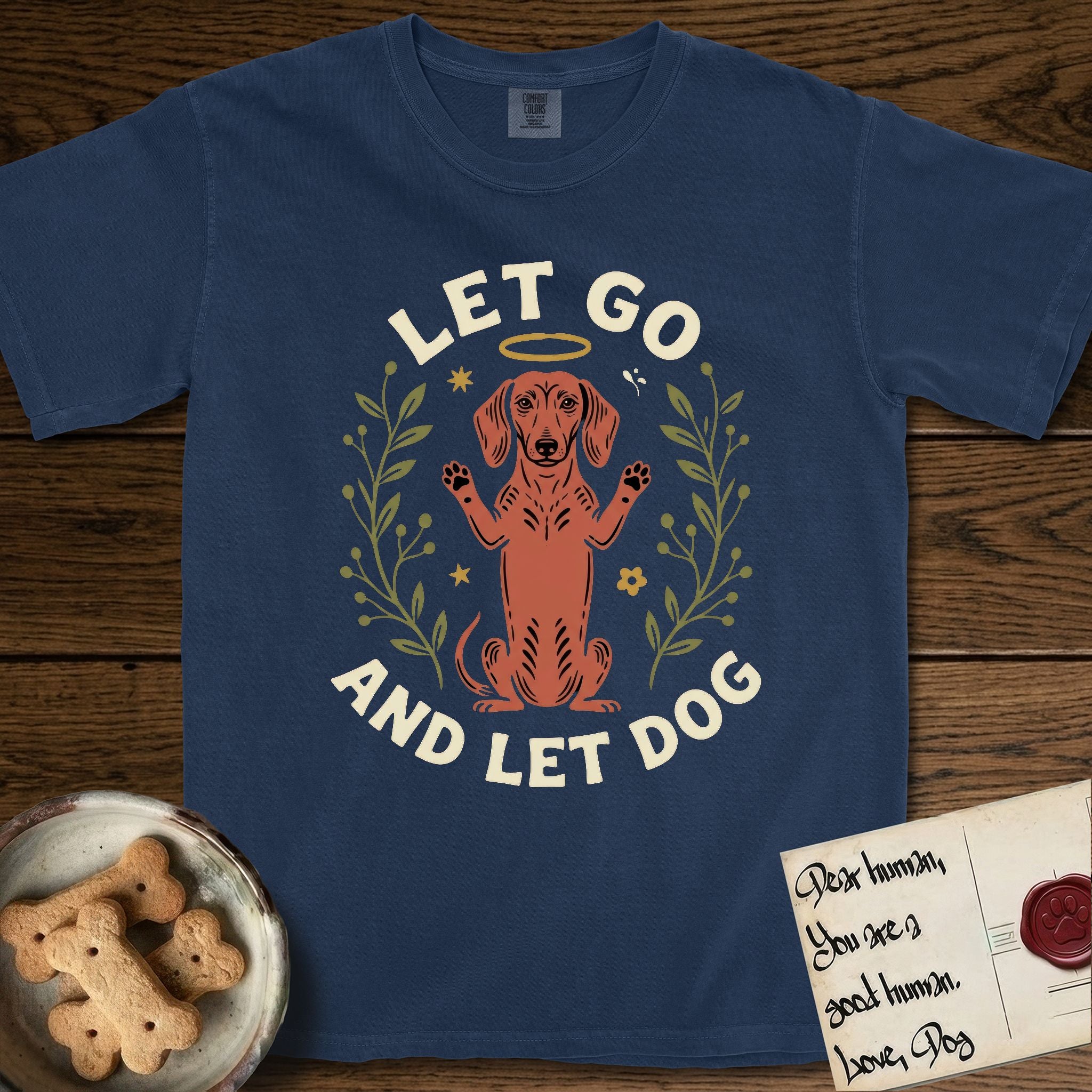 Let Go and Let Dog Dachshund T-Shirt flat lay in True Navy, featuring a folk-art botanical illustration of a haloed Dachshund sitting up with front paws raised.