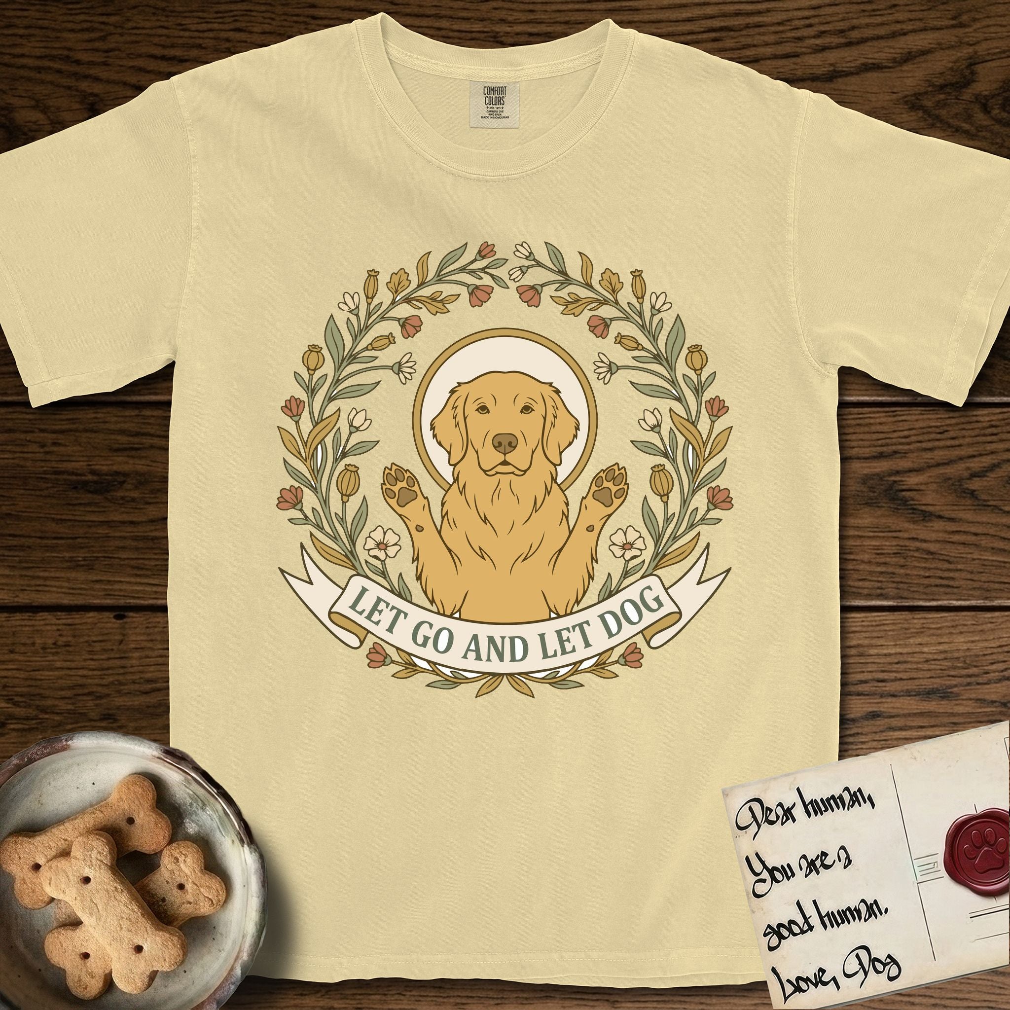 Let Go and Let Dog Golden Retriever T-Shirt flat lay in Butter, featuring an Art Nouveau wildflower wreath around a haloed Golden Retriever with front paws raised.