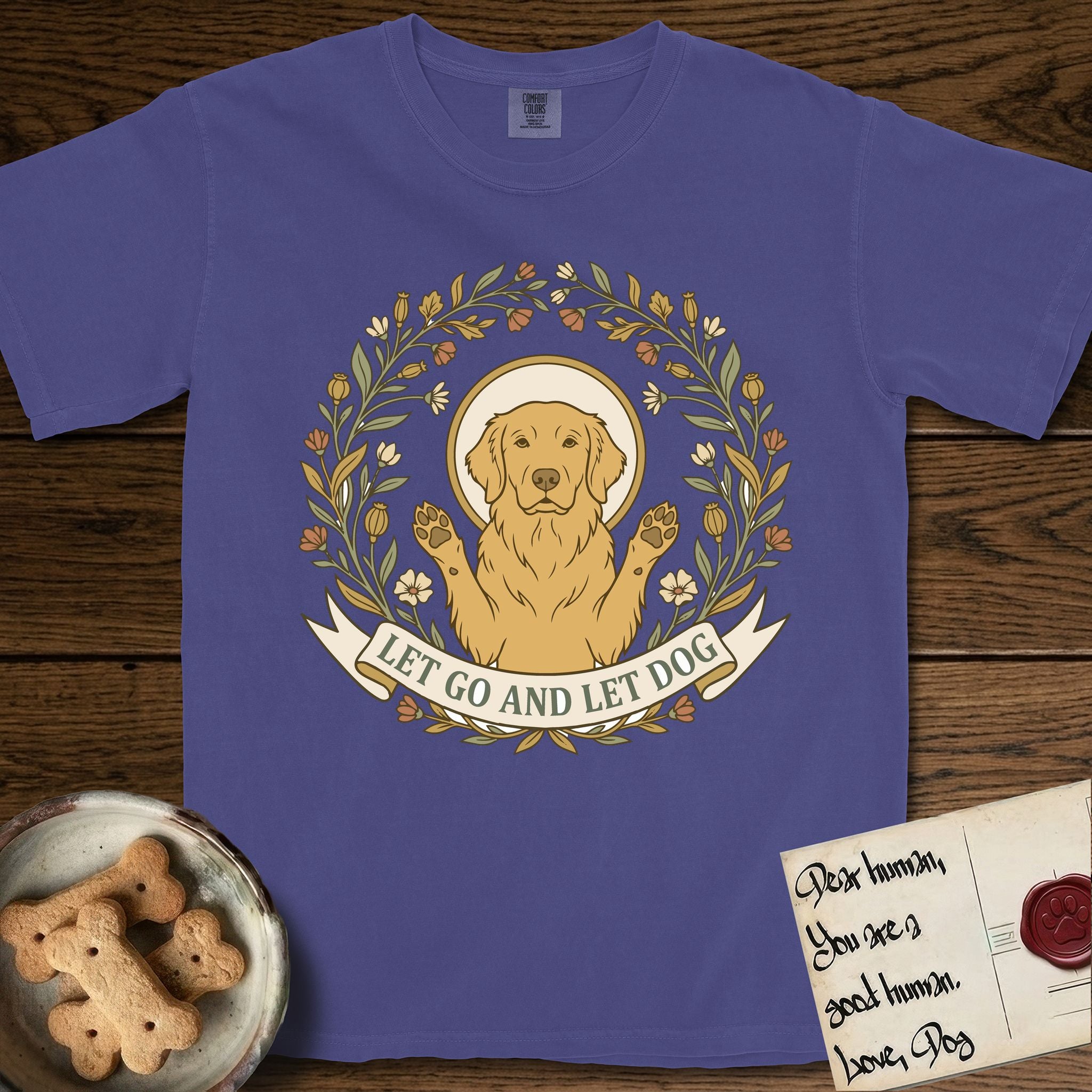 Let Go and Let Dog Golden Retriever T-Shirt flat lay in Grape, featuring an Art Nouveau wildflower wreath around a haloed Golden Retriever with front paws raised.