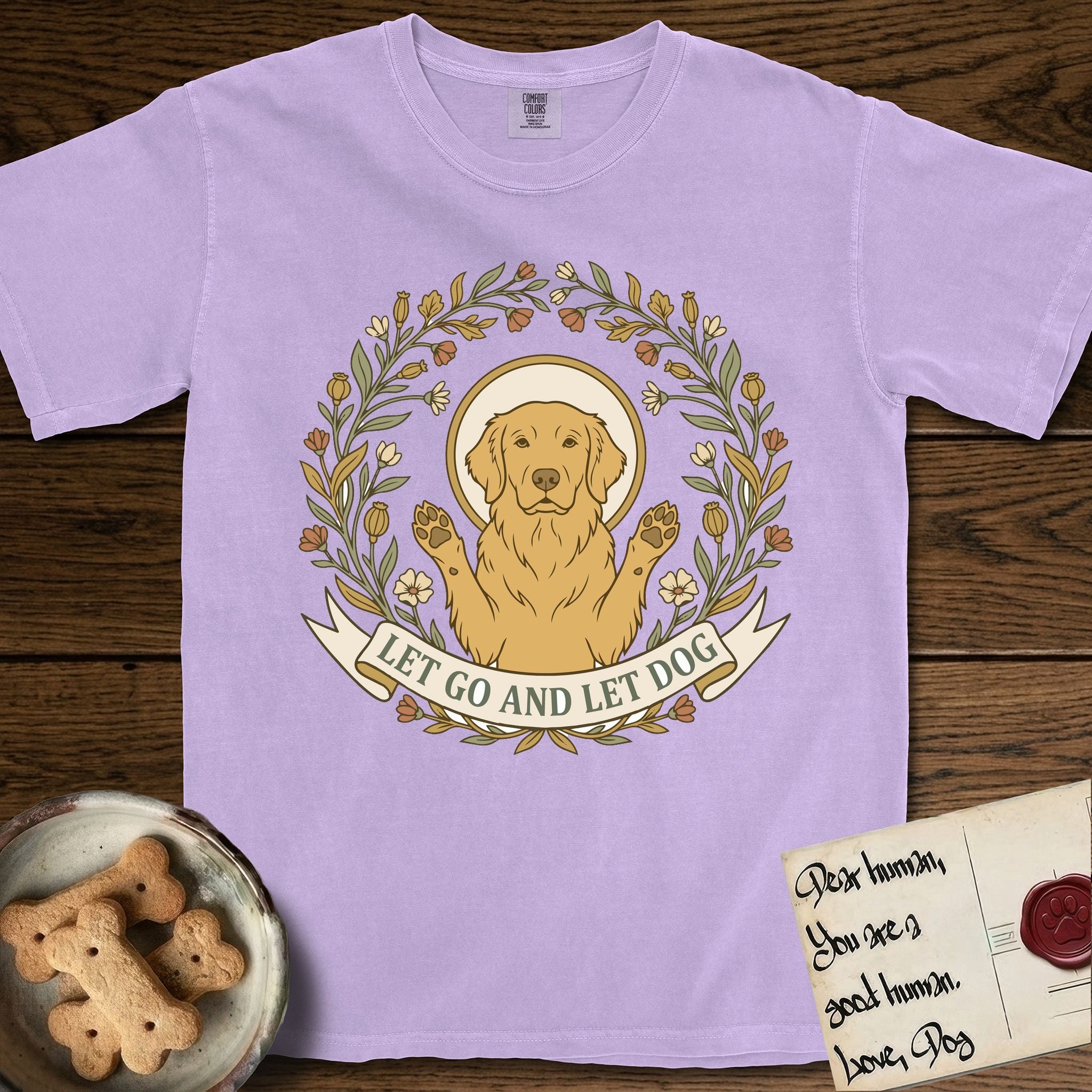 Let Go and Let Dog Golden Retriever T-Shirt flat lay in Orchid, featuring an Art Nouveau wildflower wreath around a haloed Golden Retriever with front paws raised.