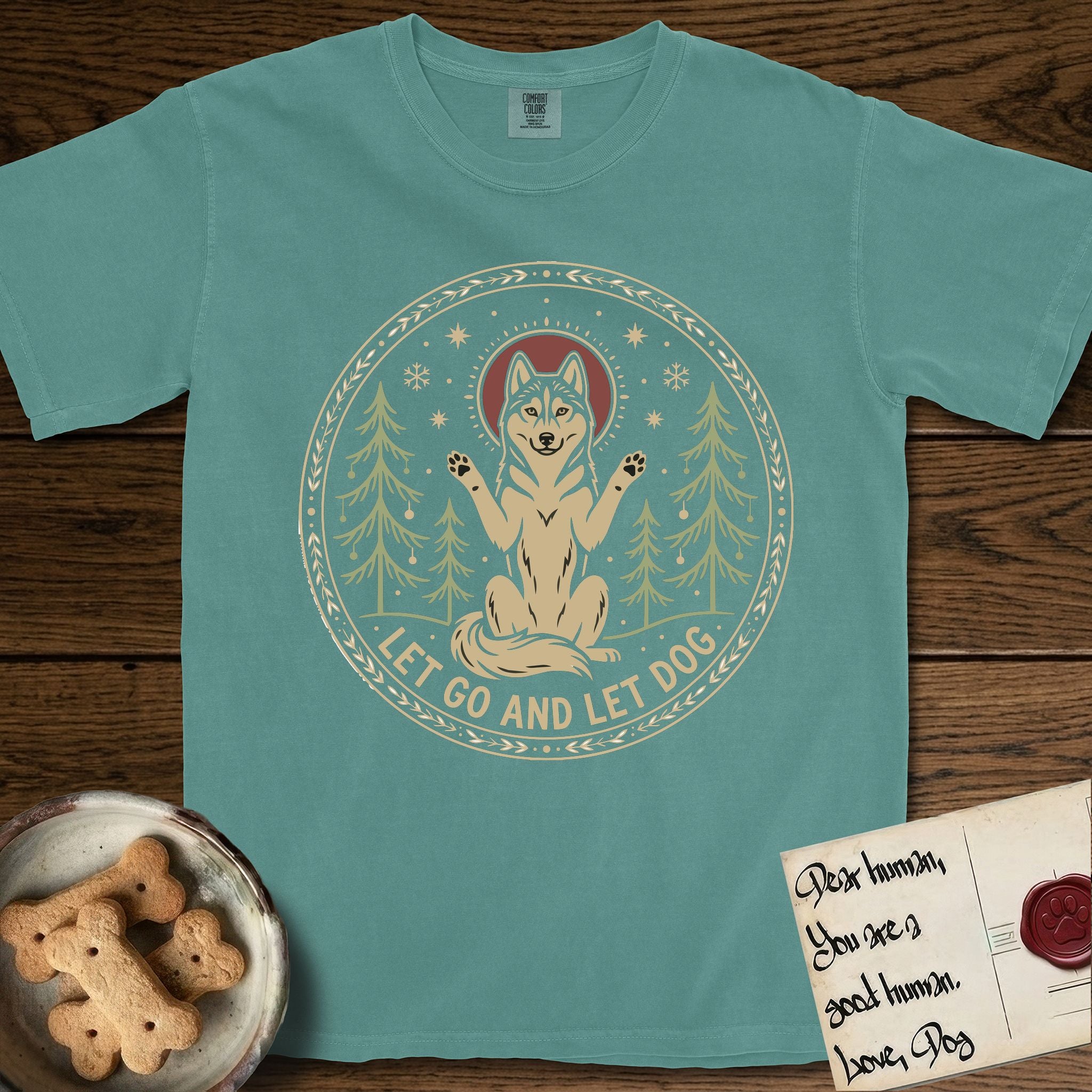 Let Go and Let Dog Husky T-Shirt flat lay in Blue Spruce, featuring a winter woodland seal of a haloed Husky sitting with paws raised among pine trees.
