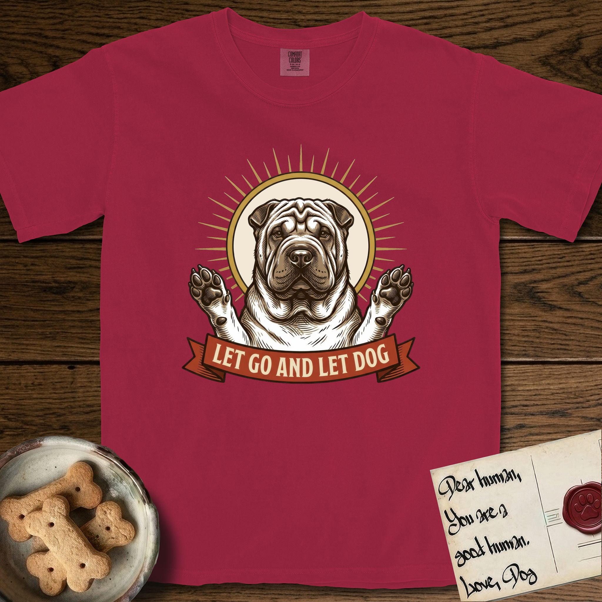 Let Go and Let Dog Shar-Pei T-Shirt flat lay in Chili, featuring an engraved woodcut portrait of a haloed wrinkled Shar-Pei, paws raised, sunburst rays behind her and a ribbon banner below.