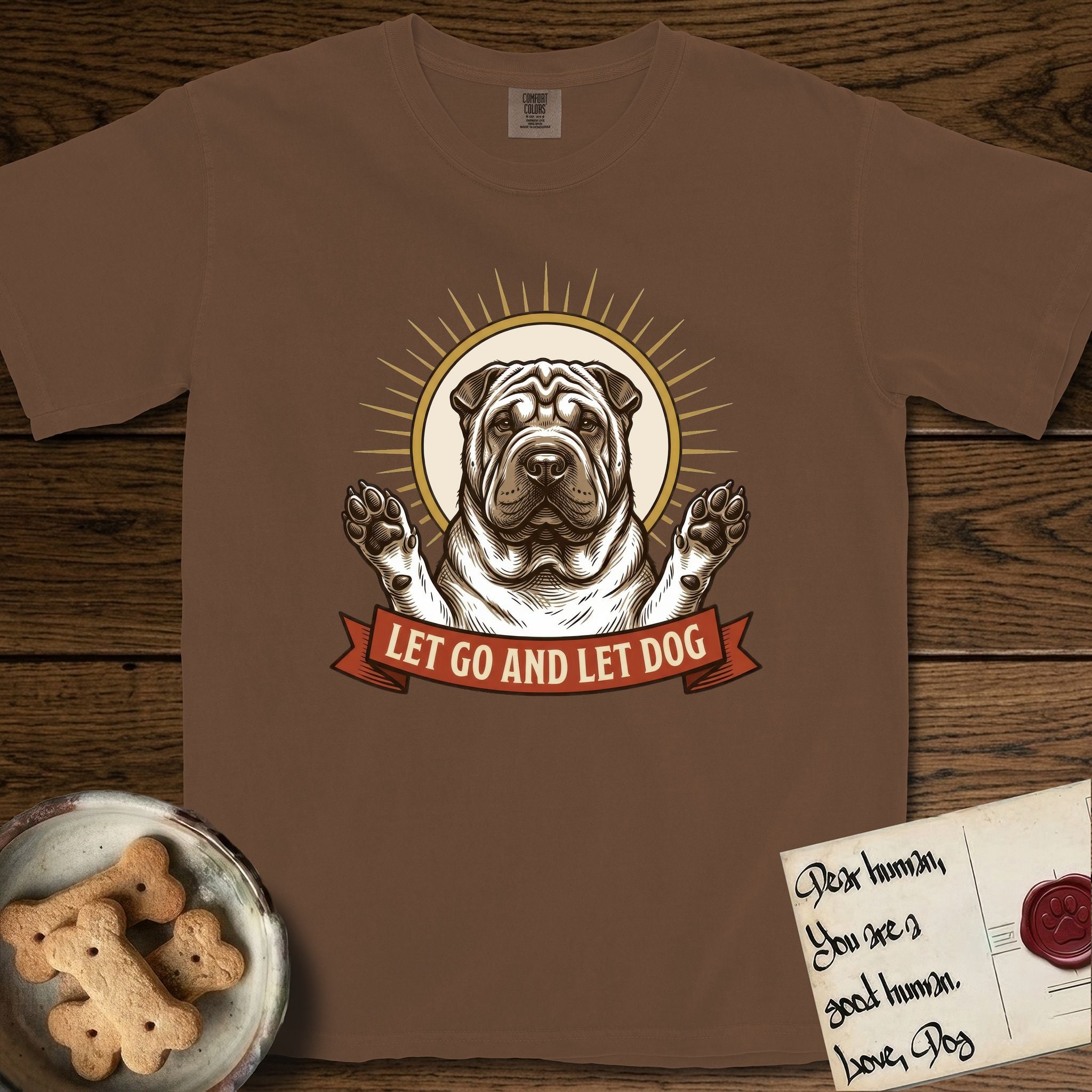 Let Go and Let Dog Shar-Pei T-Shirt flat lay in Espresso, featuring an engraved woodcut portrait of a haloed wrinkled Shar-Pei, paws raised, sunburst rays behind her and a ribbon banner below.