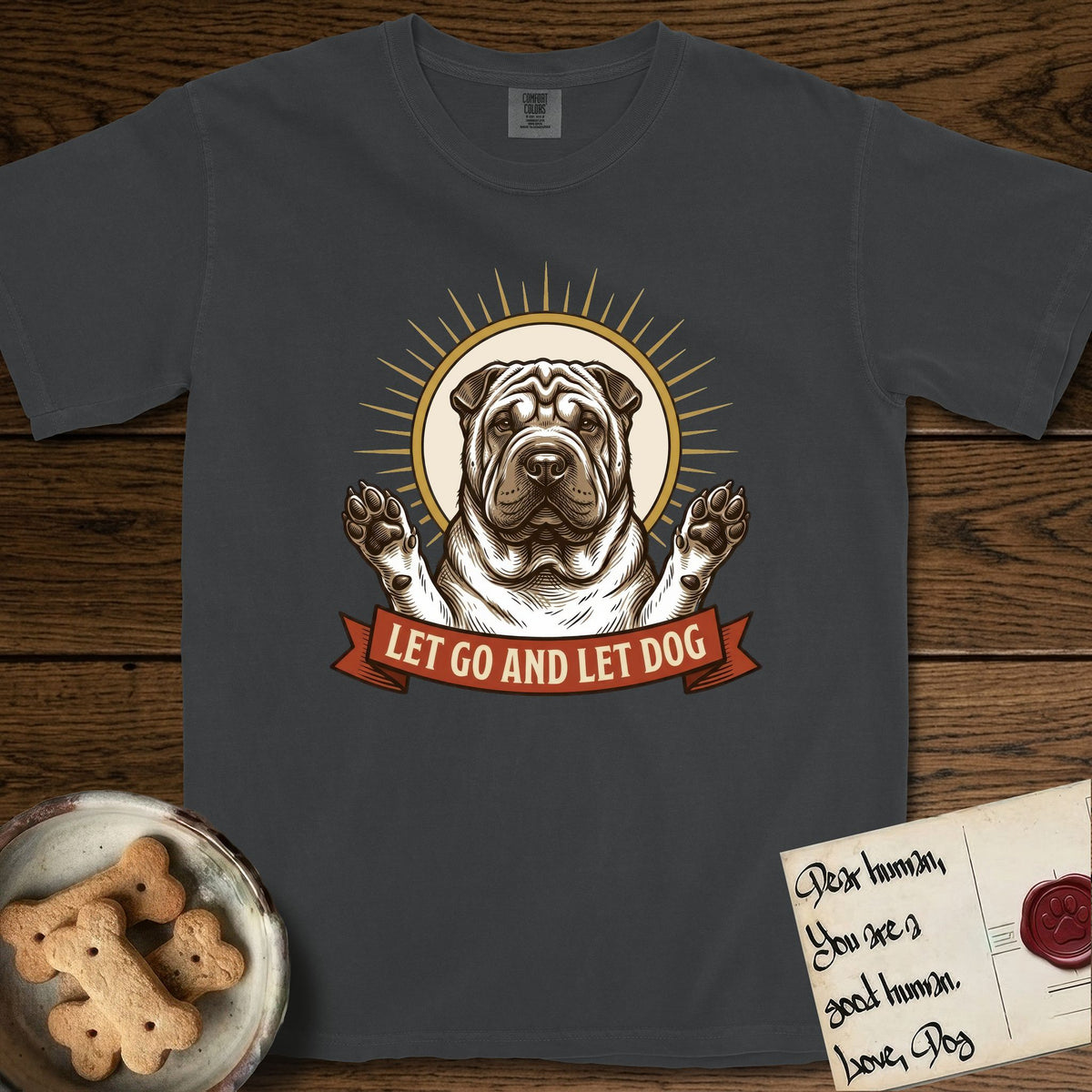 Let Go and Let Dog Shar-Pei T-Shirt flat lay in Graphite, featuring an engraved woodcut portrait of a haloed wrinkled Shar-Pei, paws raised, sunburst rays behind her and a ribbon banner below.