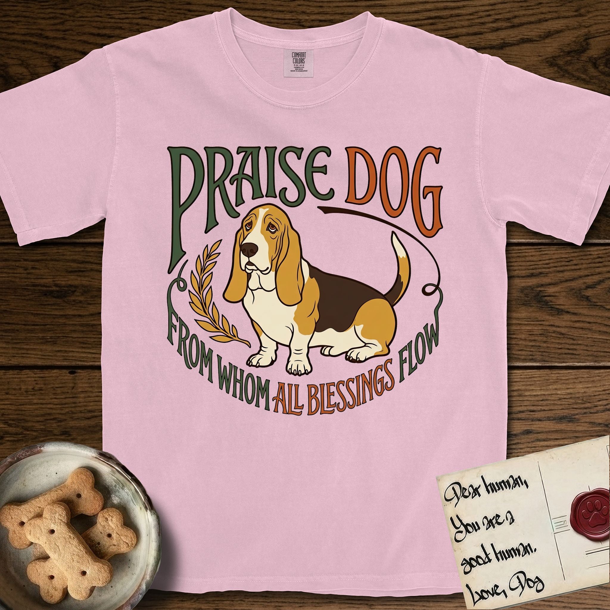 Praise Dog From Whom All Blessings Flow Basset Hound T-Shirt flat lay in Blossom, featuring an art nouveau hand-lettered design with a sad-eyed Basset Hound sitting beside a golden laurel sprig and the words Praise Dog From Whom All Blessings Flow.