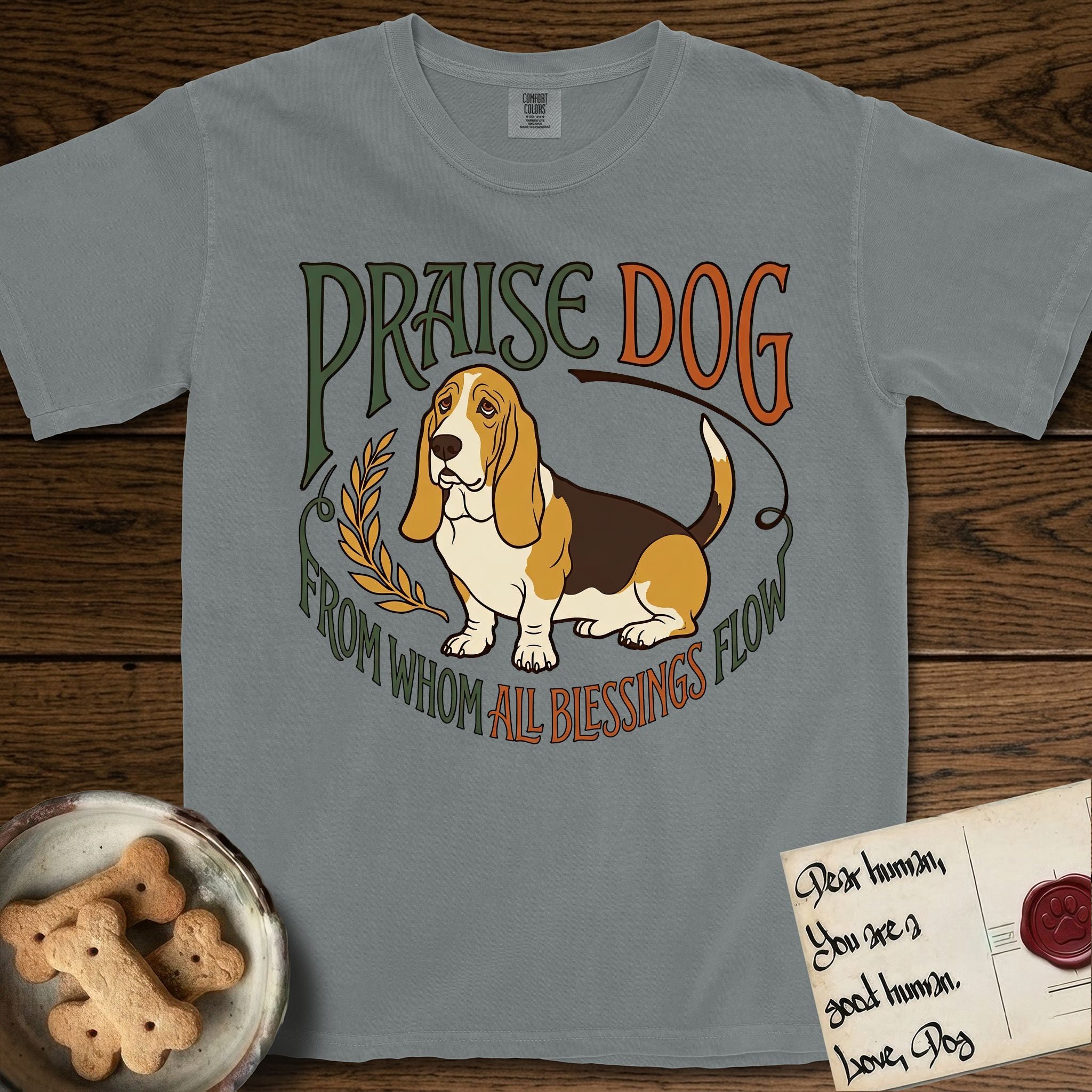 Praise Dog From Whom All Blessings Flow Basset Hound T-Shirt flat lay in Granite, featuring an art nouveau hand-lettered design with a sad-eyed Basset Hound sitting beside a golden laurel sprig and the words Praise Dog From Whom All Blessings Flow.