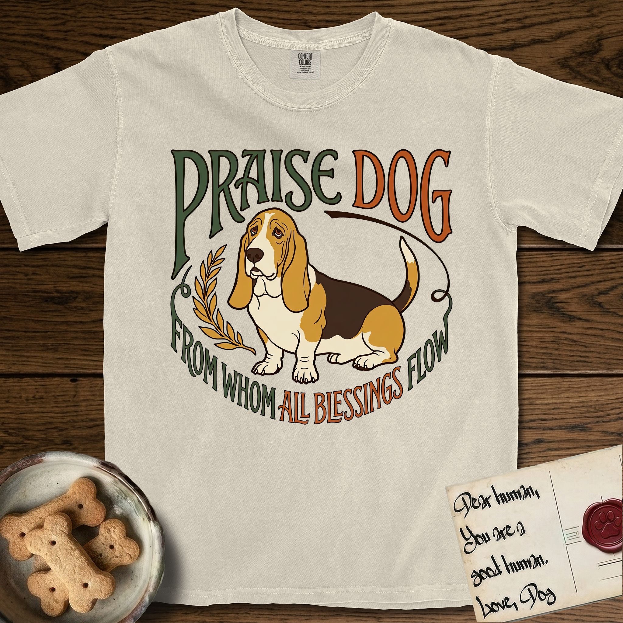 Praise Dog From Whom All Blessings Flow Basset Hound T-Shirt flat lay in Ivory, featuring an art nouveau hand-lettered design with a sad-eyed Basset Hound sitting beside a golden laurel sprig and the words Praise Dog From Whom All Blessings Flow.
