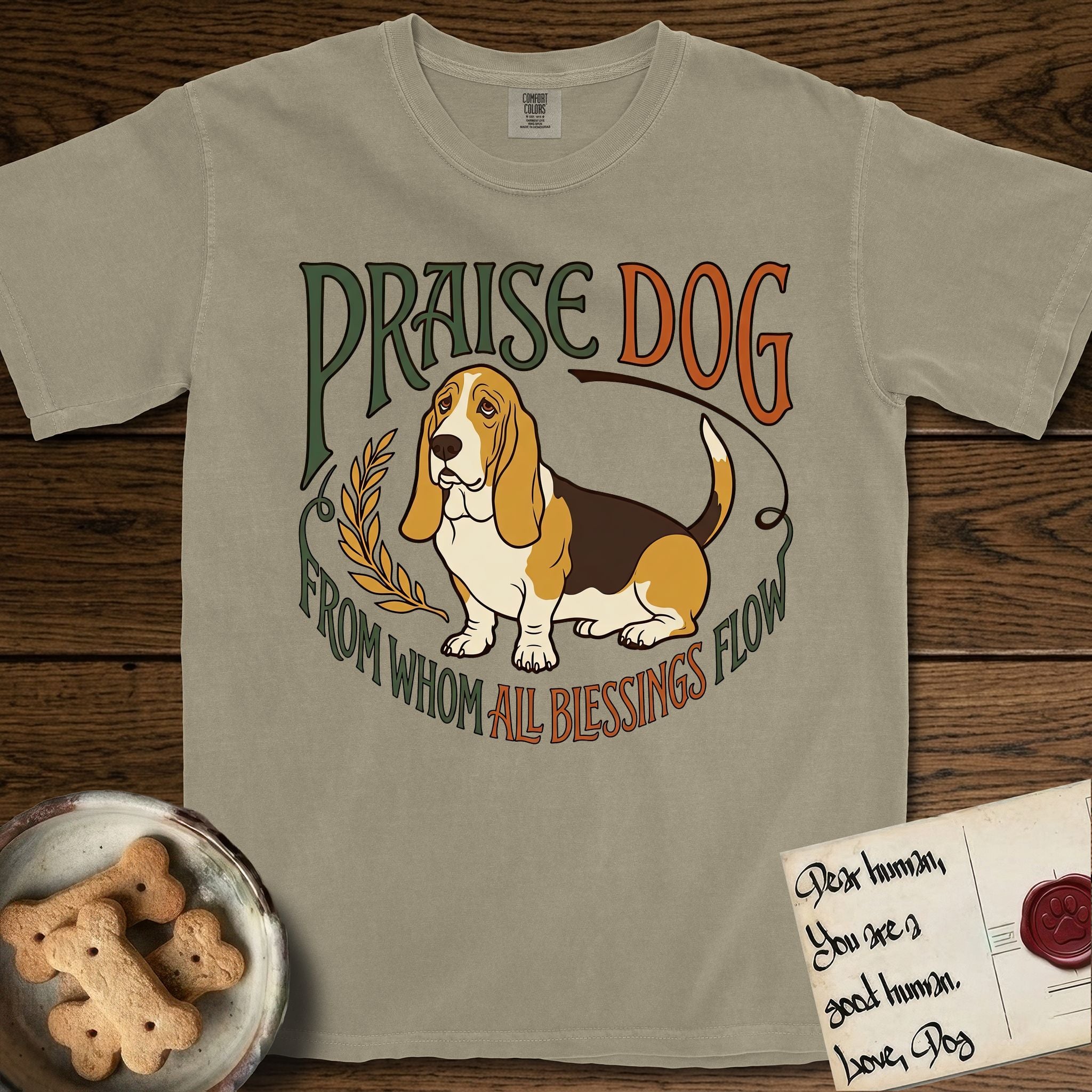 Praise Dog From Whom All Blessings Flow Basset Hound T-Shirt flat lay in Khaki, featuring an art nouveau hand-lettered design with a sad-eyed Basset Hound sitting beside a golden laurel sprig and the words Praise Dog From Whom All Blessings Flow.