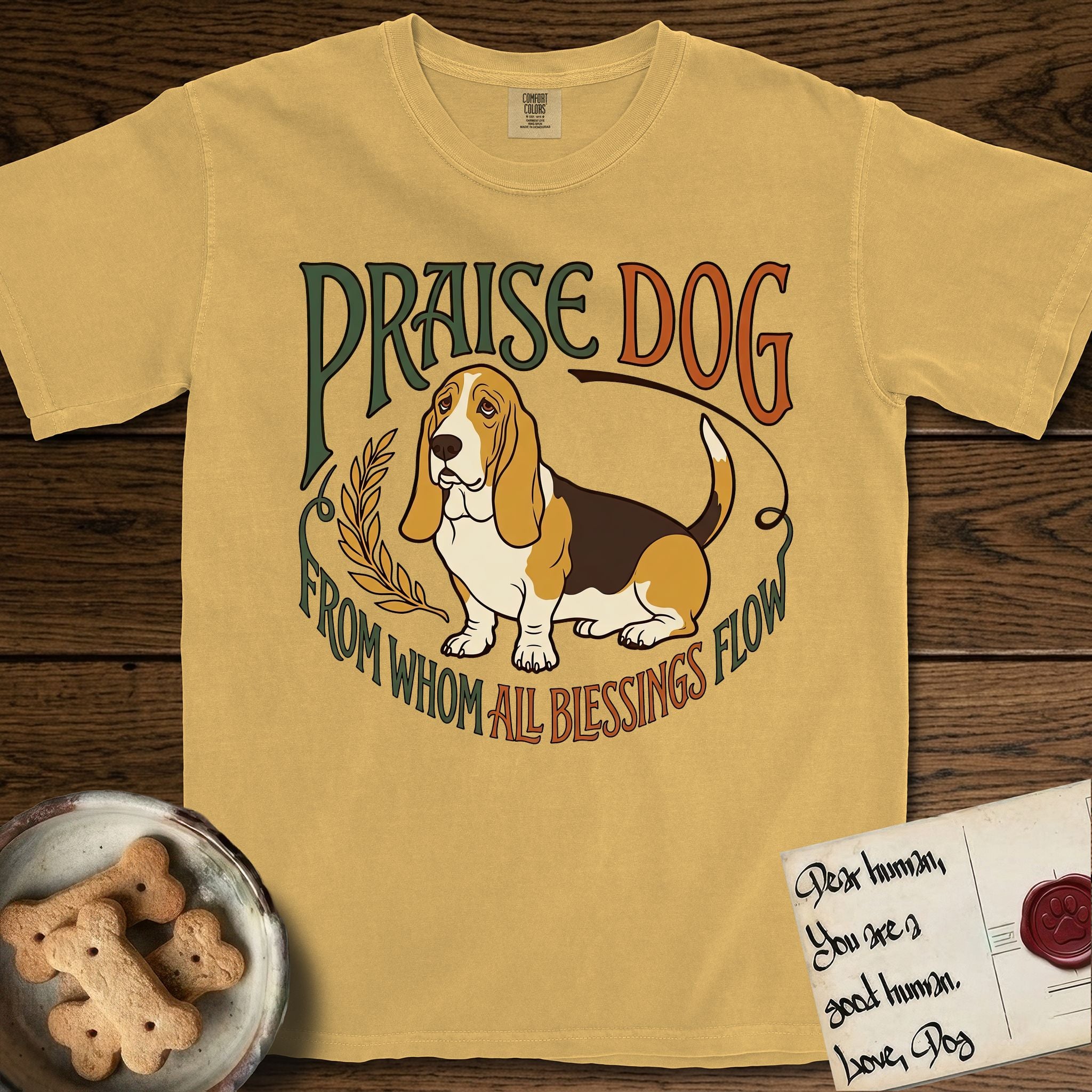 Praise Dog From Whom All Blessings Flow Basset Hound T-Shirt flat lay in Mustard, featuring an art nouveau hand-lettered design with a sad-eyed Basset Hound sitting beside a golden laurel sprig and the words Praise Dog From Whom All Blessings Flow.