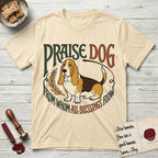 Praise Dog From Whom All Blessings Flow Basset Hound T-Shirt flat lay in Natural, featuring an art nouveau hand-lettered design with a sad-eyed Basset Hound sitting beside a golden laurel sprig and the words Praise Dog From Whom All Blessings Flow.