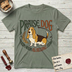 Praise Dog From Whom All Blessings Flow Basset Hound T-Shirt flat lay in Sage, featuring an art nouveau hand-lettered design with a sad-eyed Basset Hound sitting beside a golden laurel sprig and the words Praise Dog From Whom All Blessings Flow.